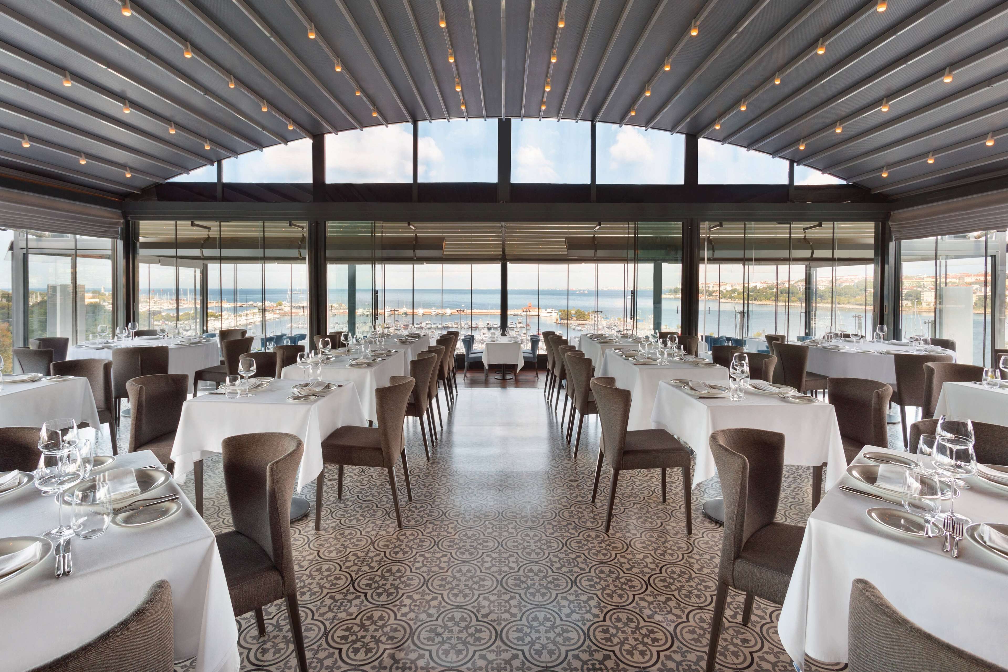 A restaurant with a long table layout, large windows, and a view of the sea.
