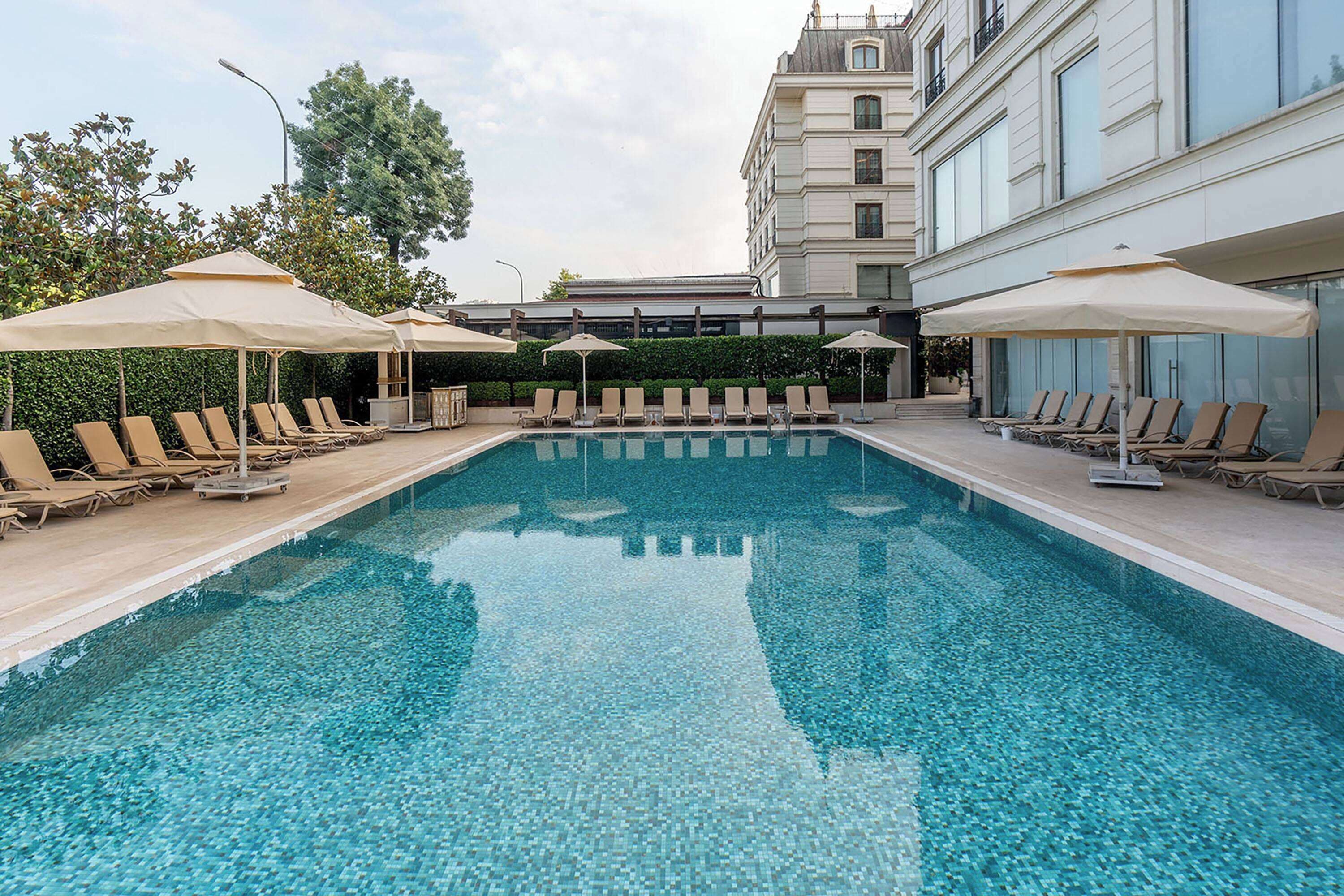 A swimming pool with lounge chairs and umbrellas, surrounded by a building with large windows and a balcony.