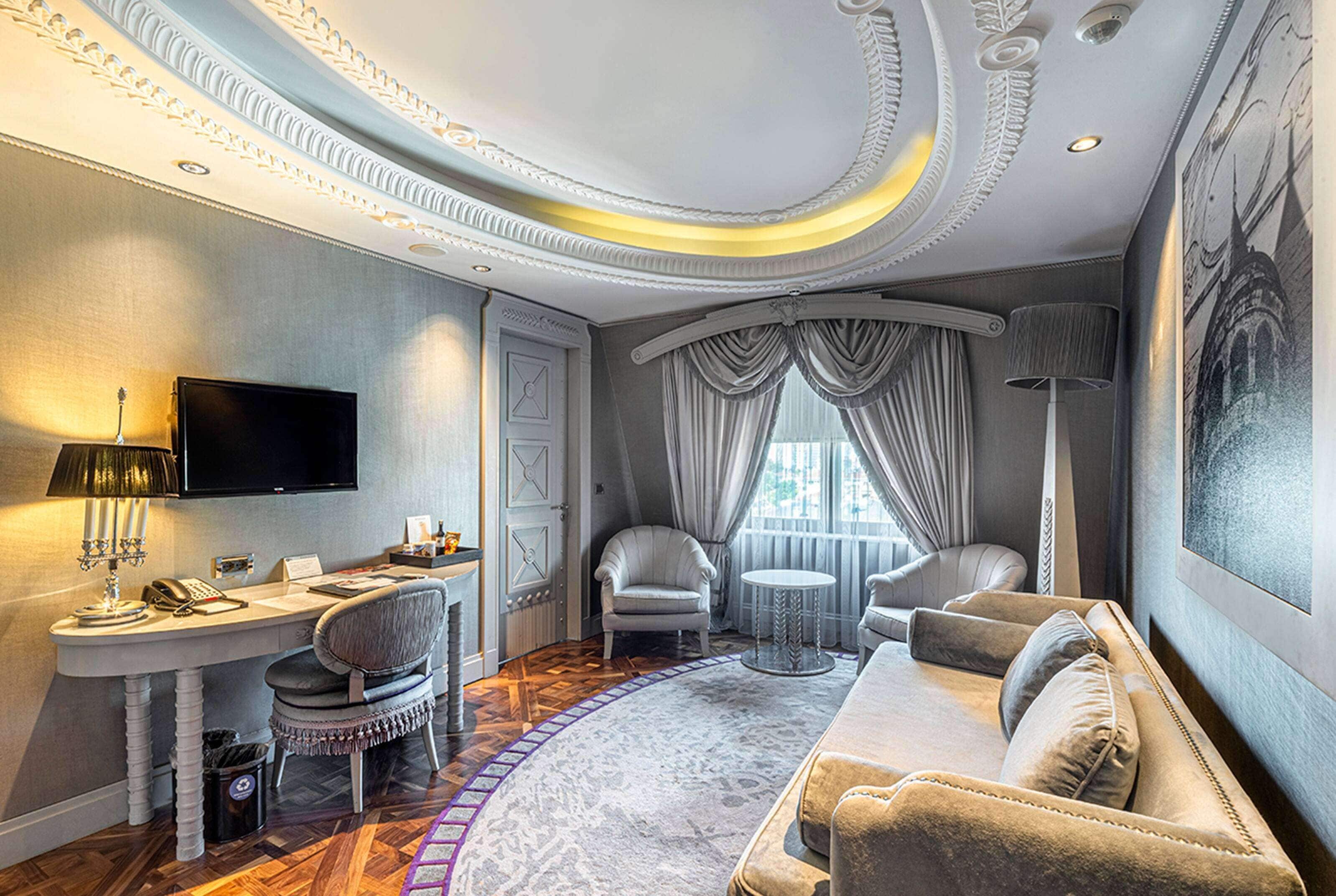 A luxurious room with a curved sofa, two armchairs, a desk with a flat-screen TV, a lamp, and a large window with curtains.