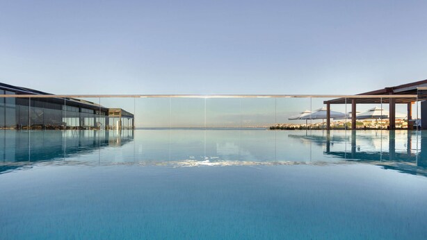 Una gran piscina cristalina con piso de vidrio, rodeada de edificios modernos y un cielo despejado.