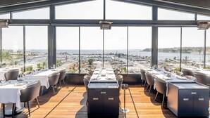 Un restaurante con una larga mesa y sillas, amplias ventanas y vistas a un puerto deportivo.