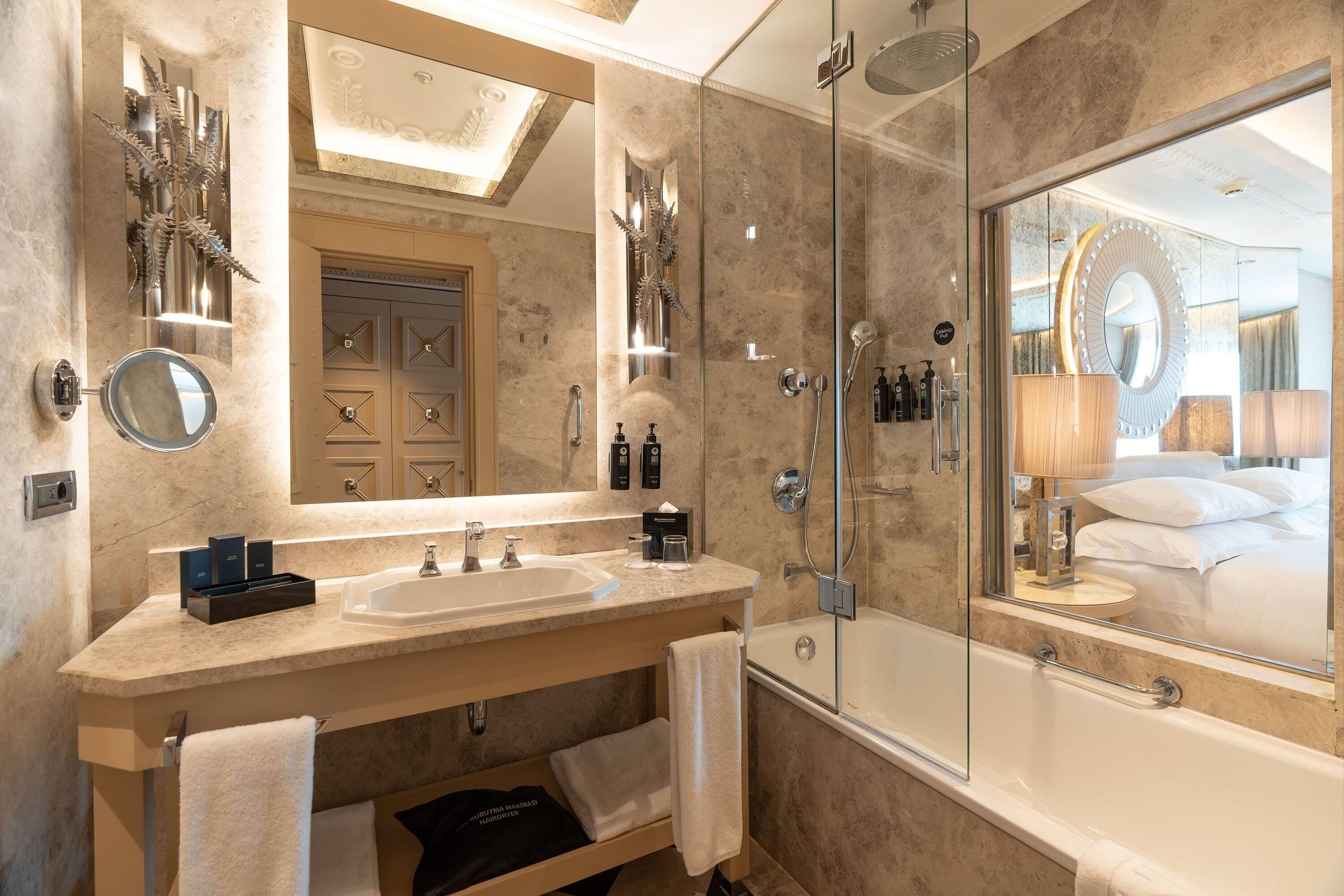 A modern bathroom with a double sink vanity, a glass-enclosed shower, and a large mirror reflecting a bedroom with a bed and pillows.