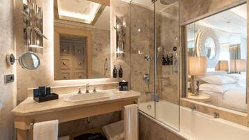 A modern bathroom with a double sink vanity, a glass-enclosed shower, and a large mirror reflecting a bedroom with a bed and pillows.