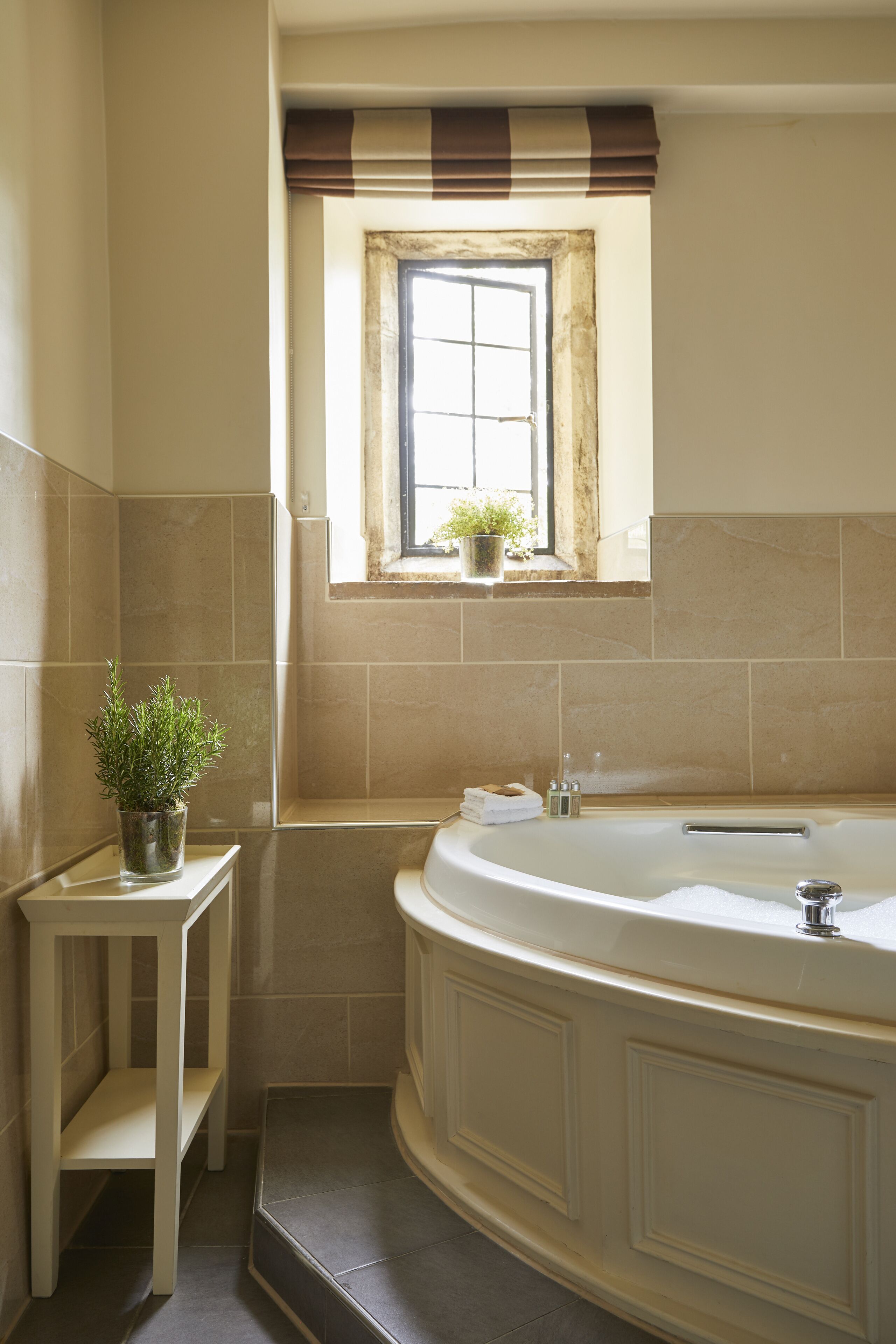 A bathroom with a large bathtub, a small window with a plant, and a shelf with toiletries.
