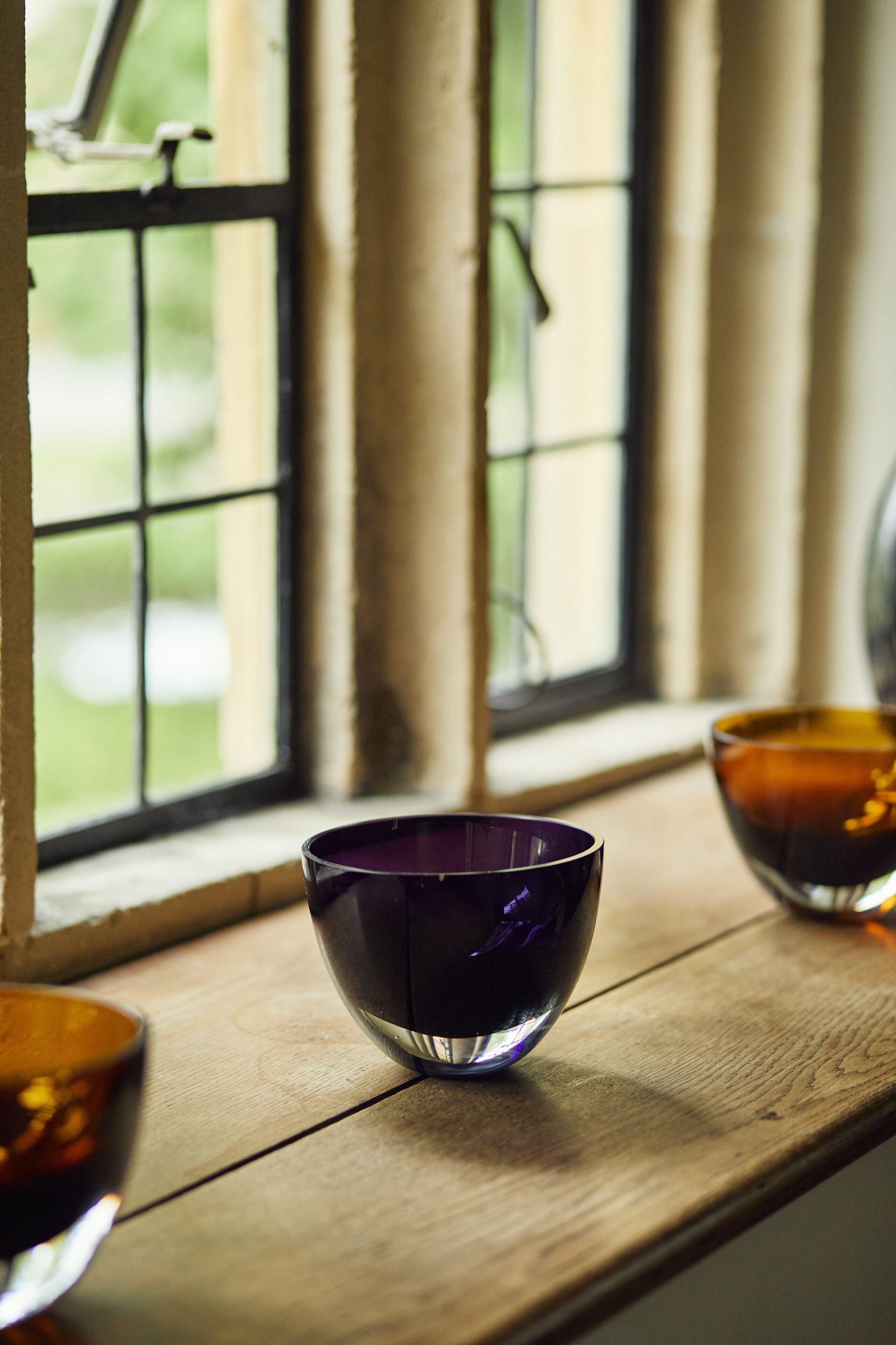 Une étagère en bois sur laquelle reposent deux bols, l’un violet et l’autre orange, près d’une fenêtre donnant sur un paysage verdoyant.