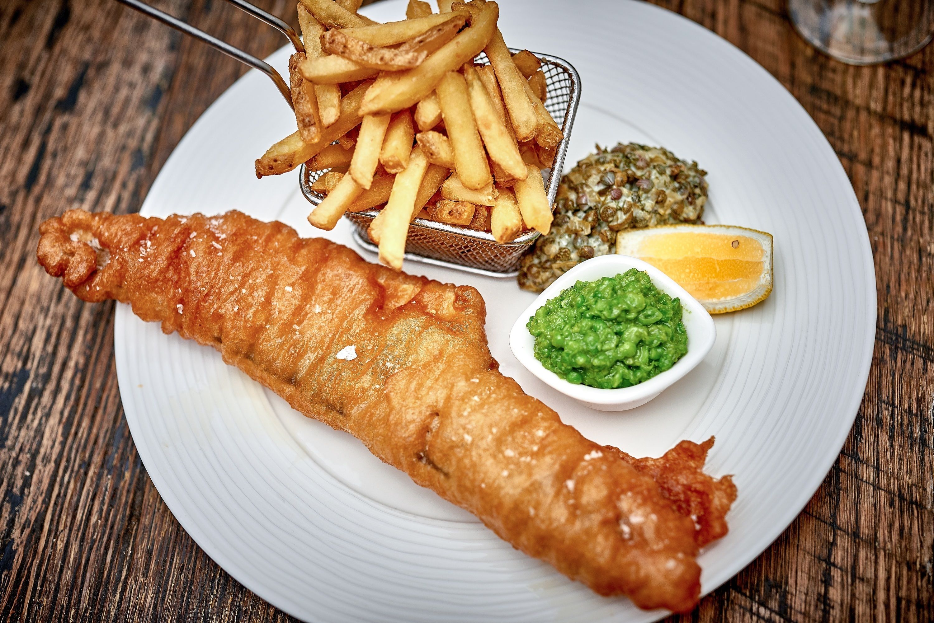 A plate with a large piece of battered fish, a basket of fries, a small bowl of green sauce, and a lemon wedge.