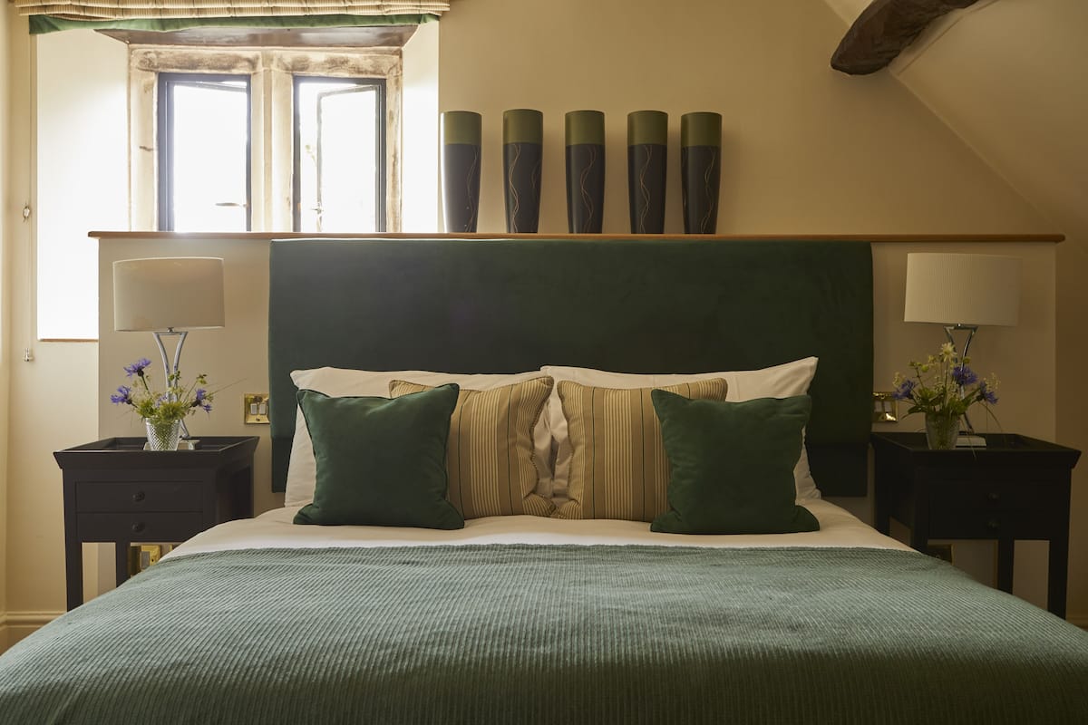 A neatly made bed with a green bedspread, two green pillows, and a striped headboard. There are two bedside tables with lamps and vases of flowers.