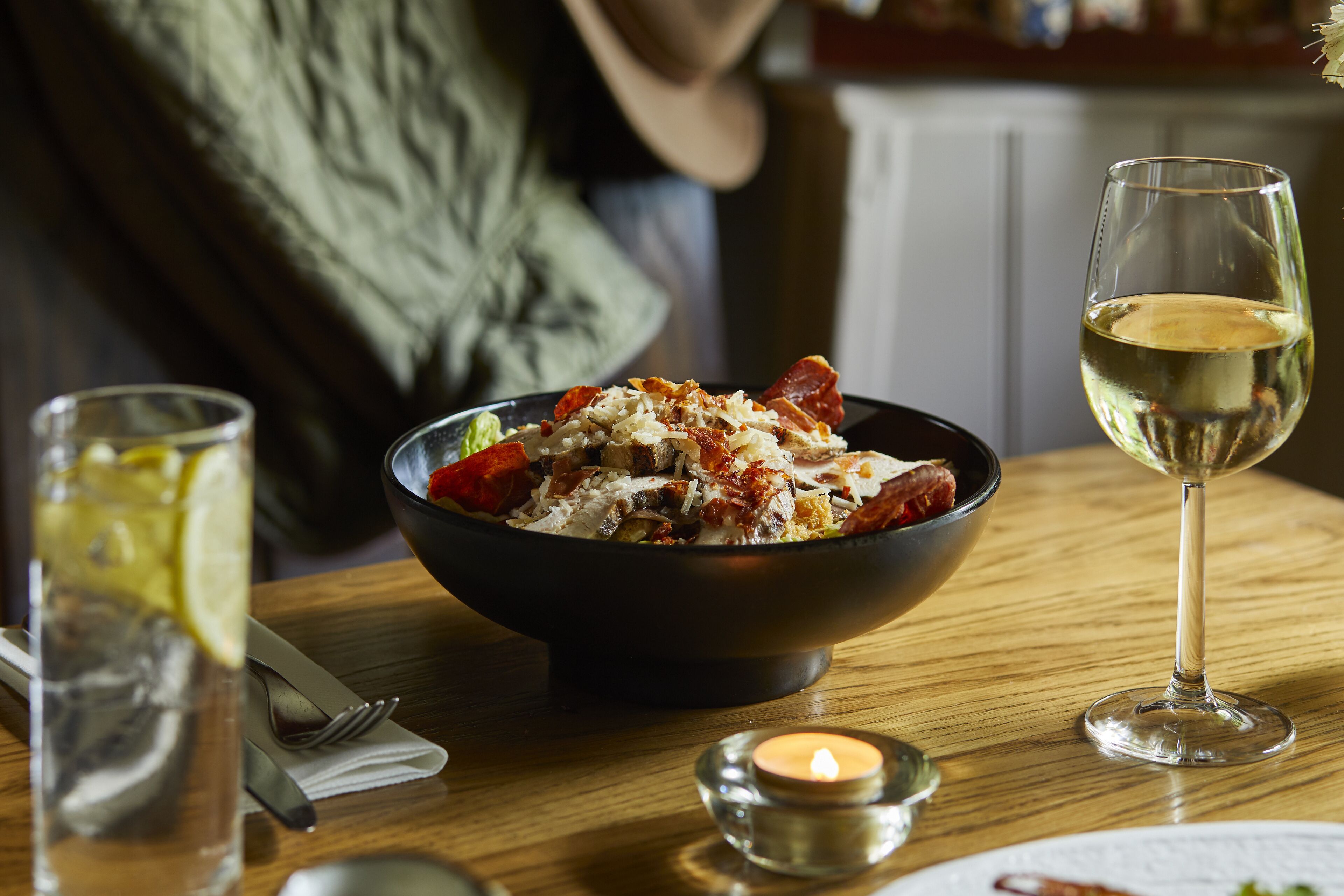 A dining table with a bowl of food, a glass of white wine, and a candle.