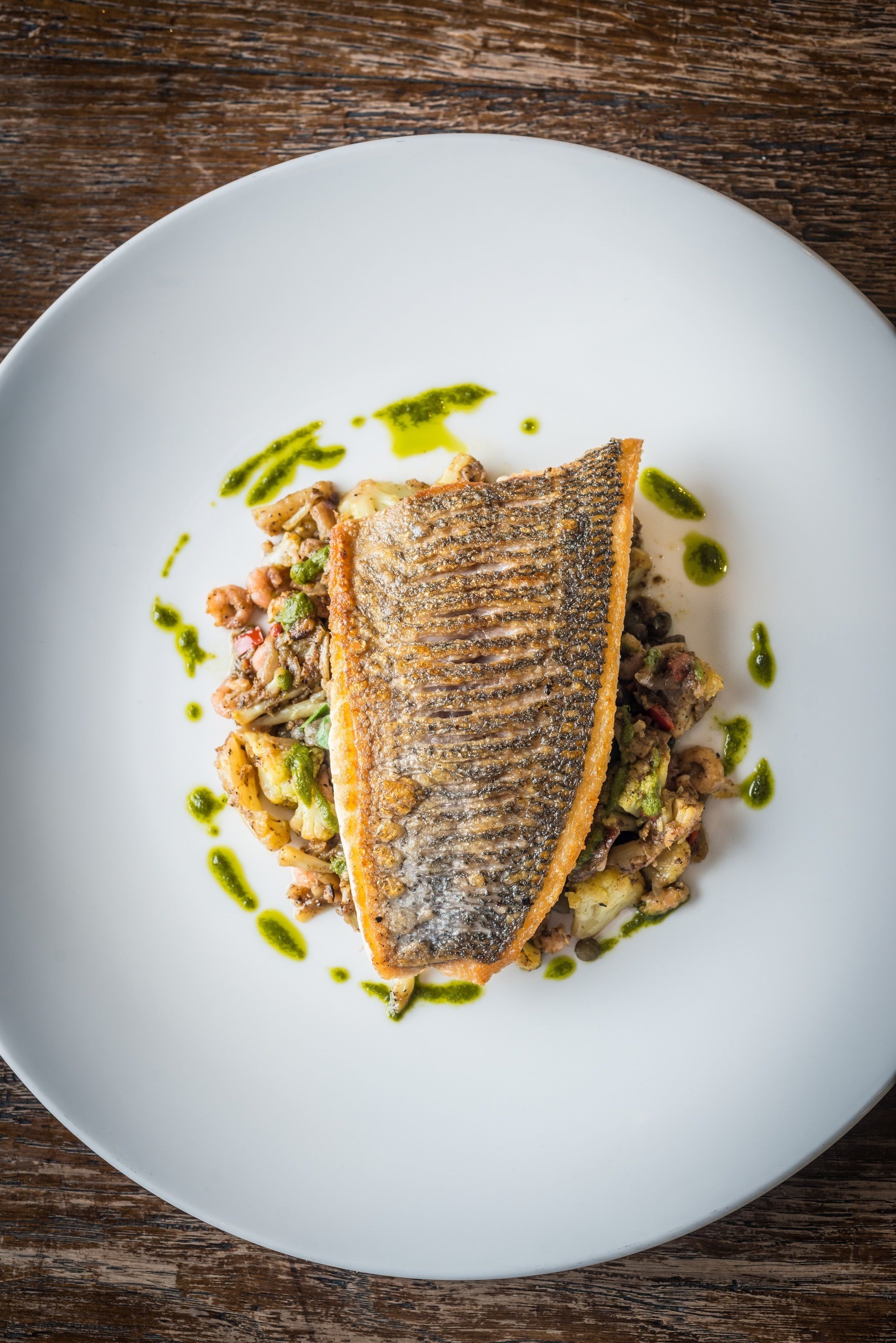 A white plate with a grilled fish, vegetables, and green sauce.