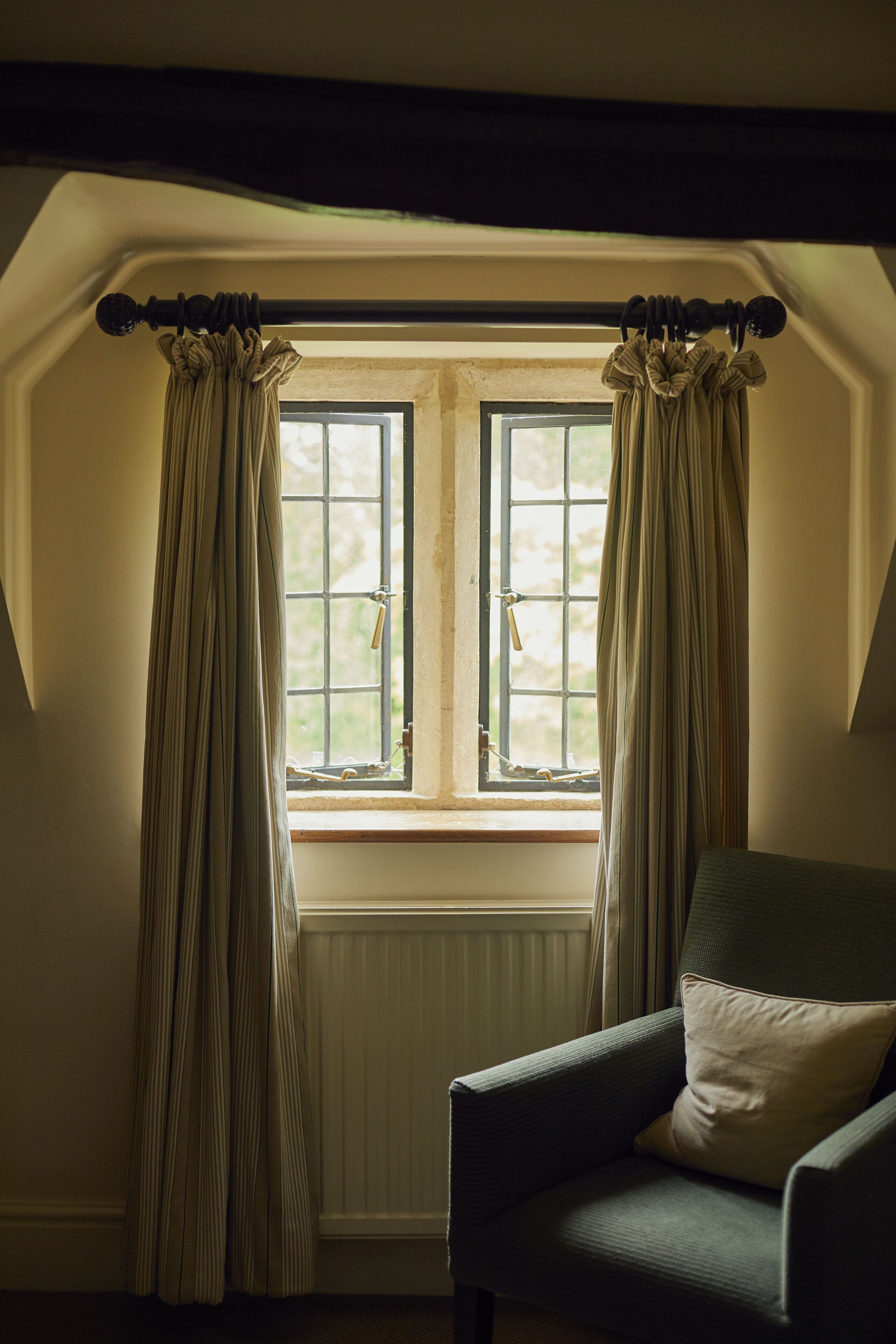 A room with a window, curtains, a radiator, and a chair with a cushion.