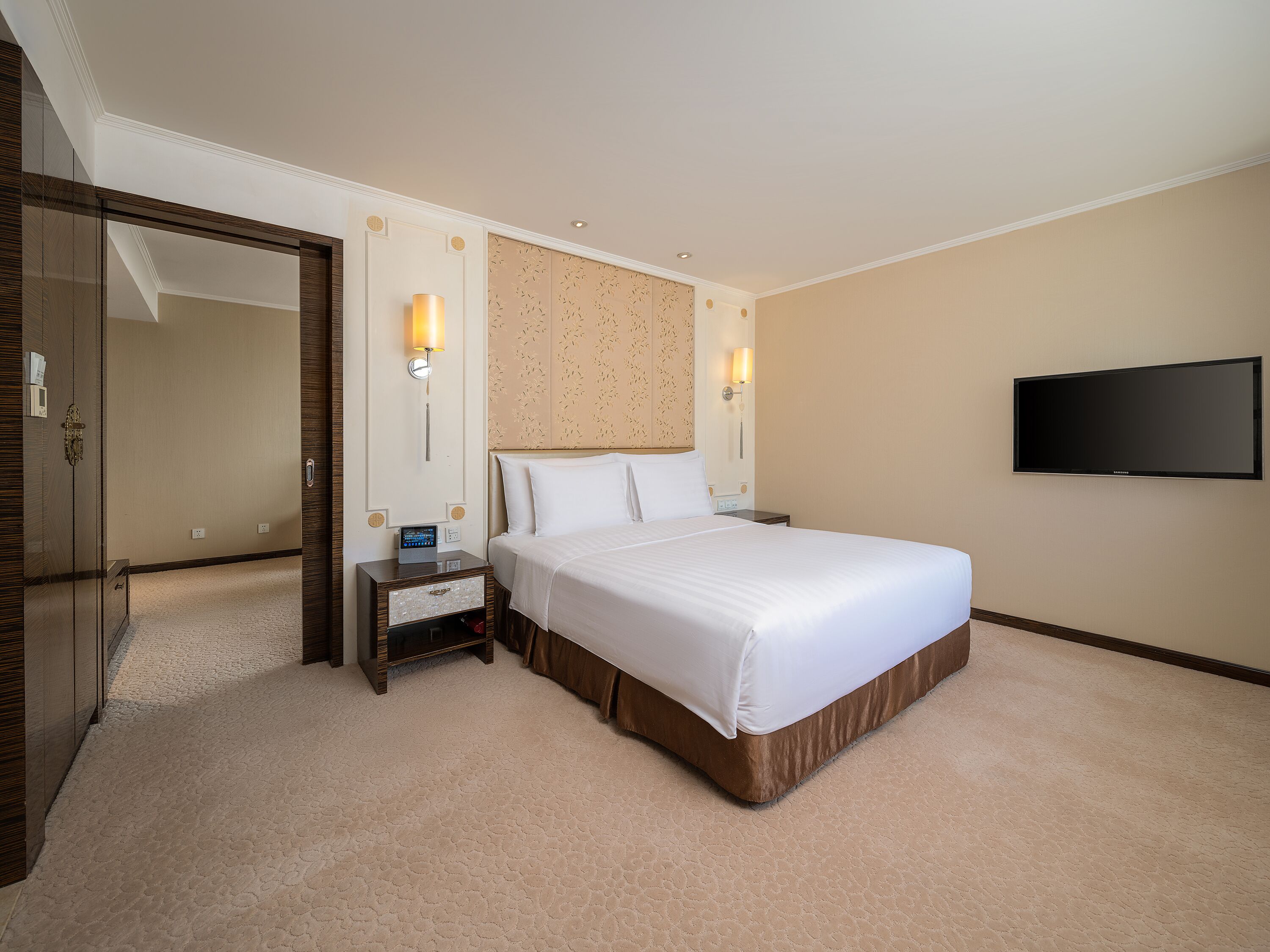 Una habitación de hotel con una cama grande, una mesita de noche, un televisor montado en la pared y una puerta que da a otra habitación.