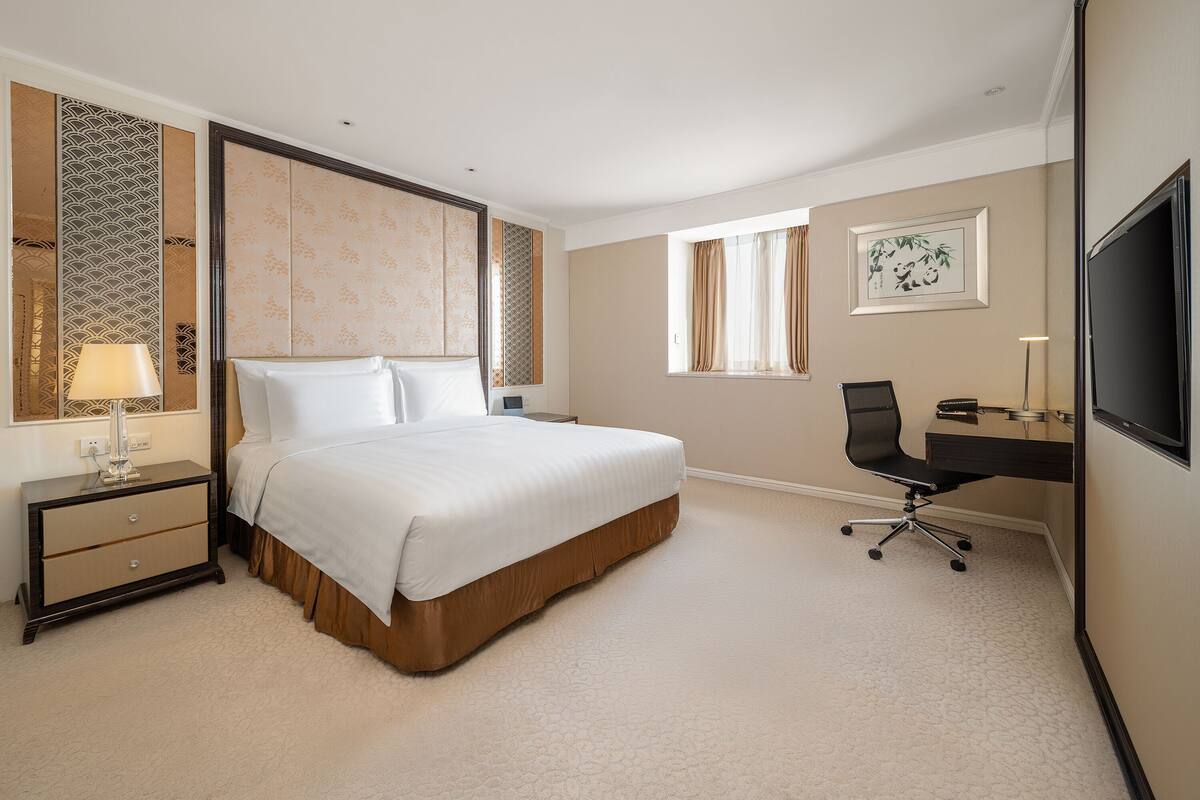 A hotel room with a large bed, a desk with a chair, a TV, and a window with curtains.