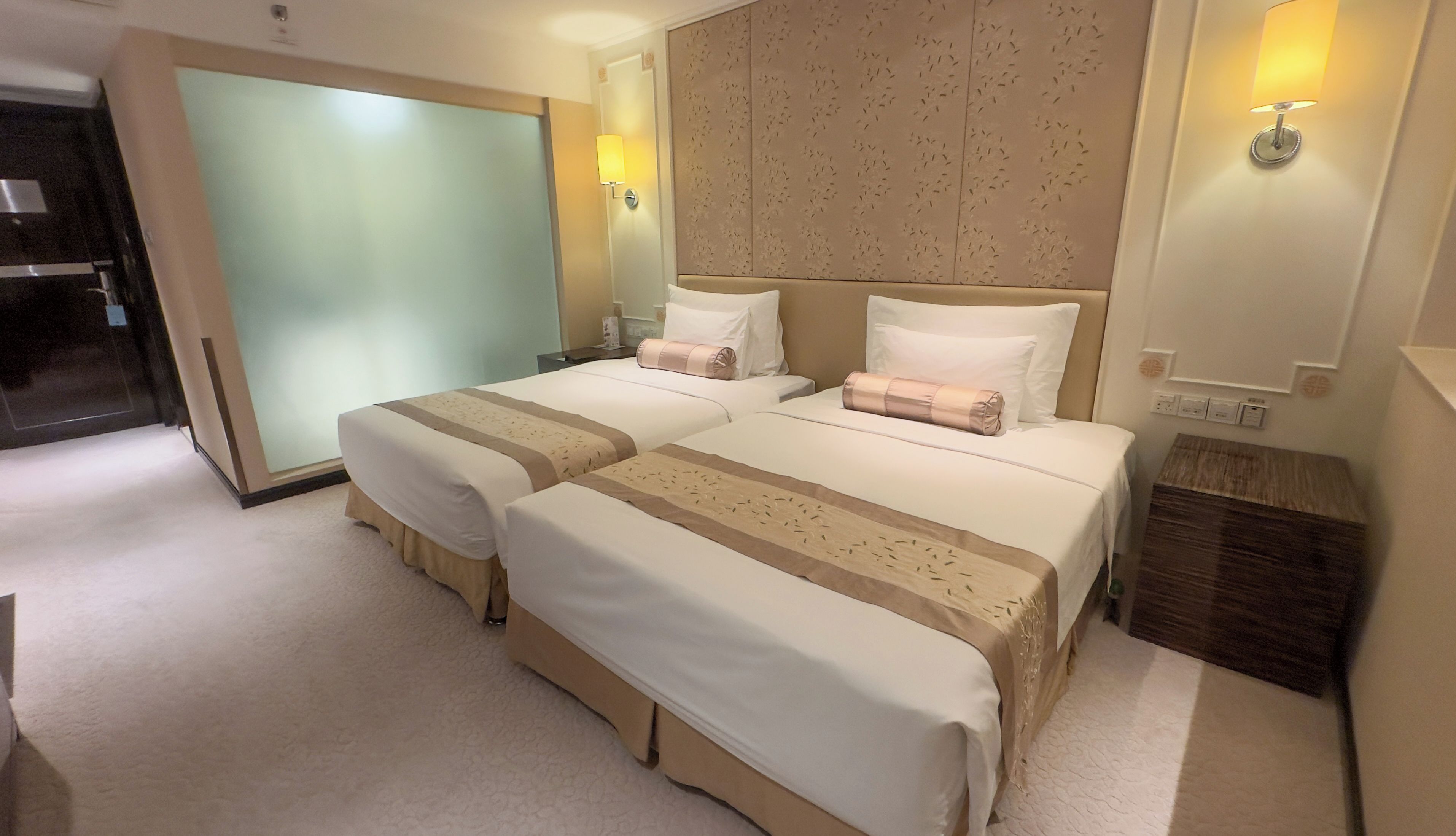 A hotel room with two beds, a wardrobe, a bedside table, and a wall-mounted lamp.
