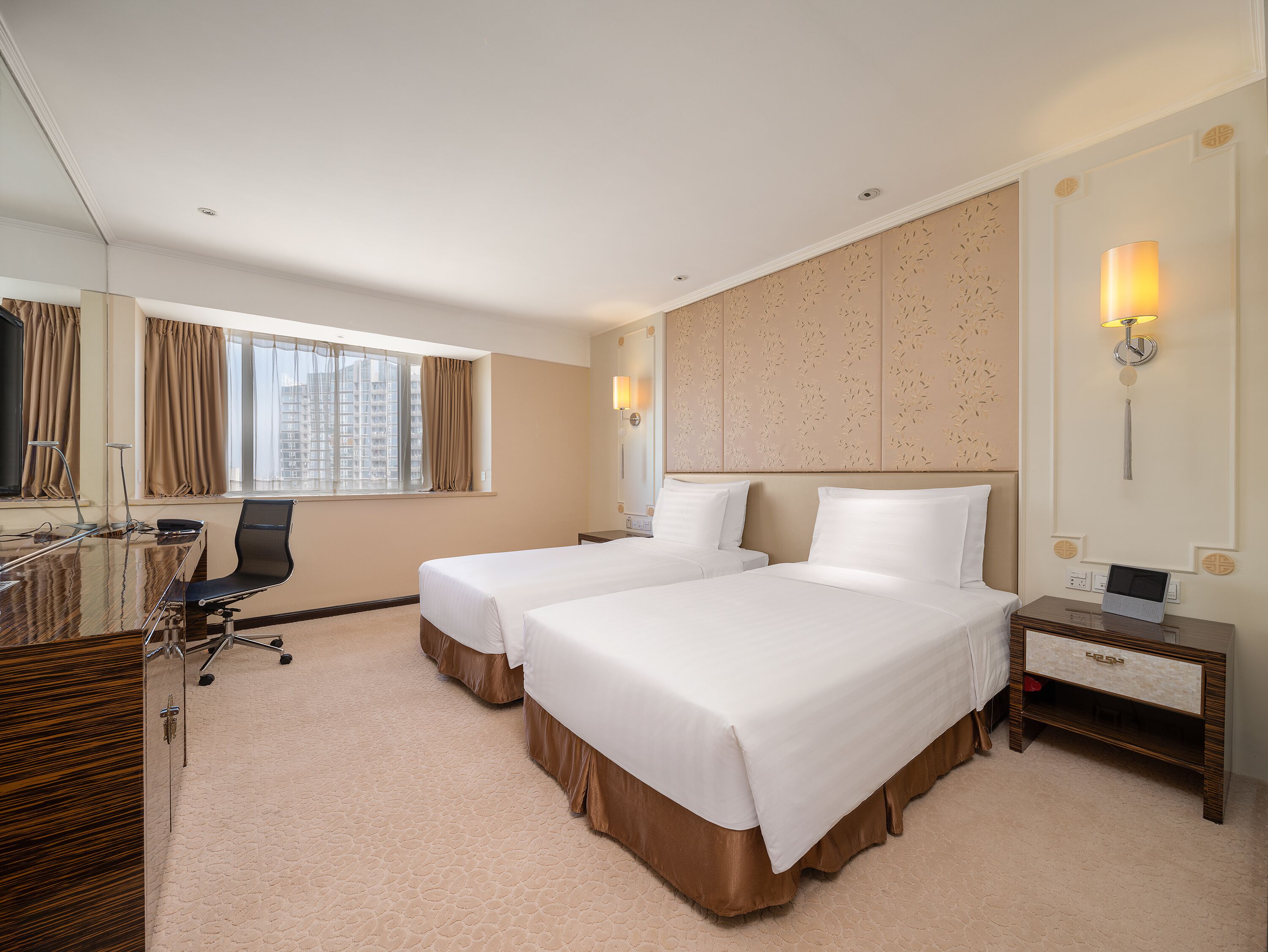 A hotel room with two beds, a desk, a chair, a lamp, and a view of the city.