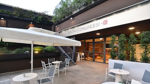An outdoor patio area with white tables and chairs, a covered seating area, and a staircase leading to a building with a sign reading "CENTRO CONGRESSI".