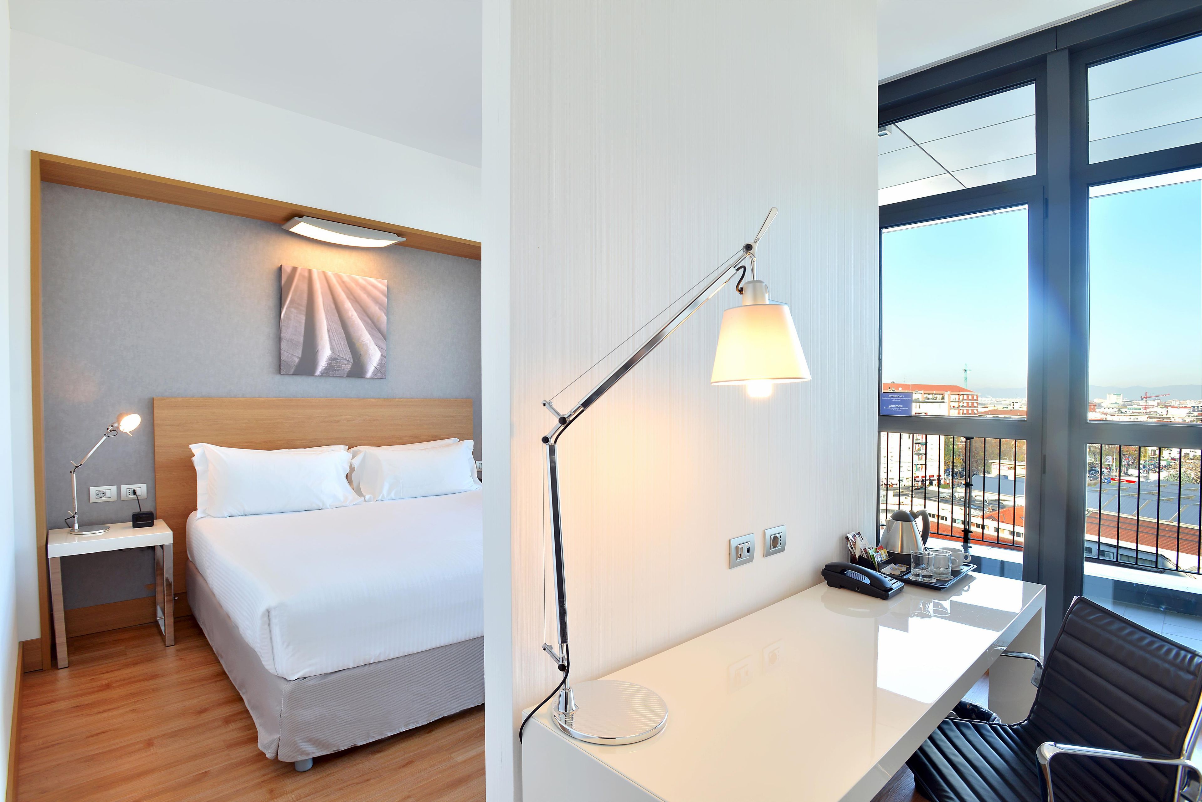 Quarto de hotel com cama, mesa de cabeceira, luminária e escrivaninha com cadeira.