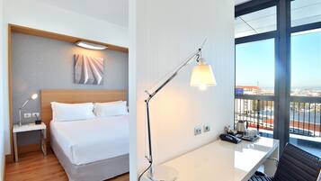 Quarto de hotel com cama, mesa de cabeceira, luminária e escrivaninha com cadeira.