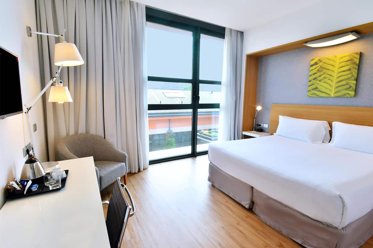 A hotel room with a large bed, a desk with a lamp, a chair, and a window with curtains.