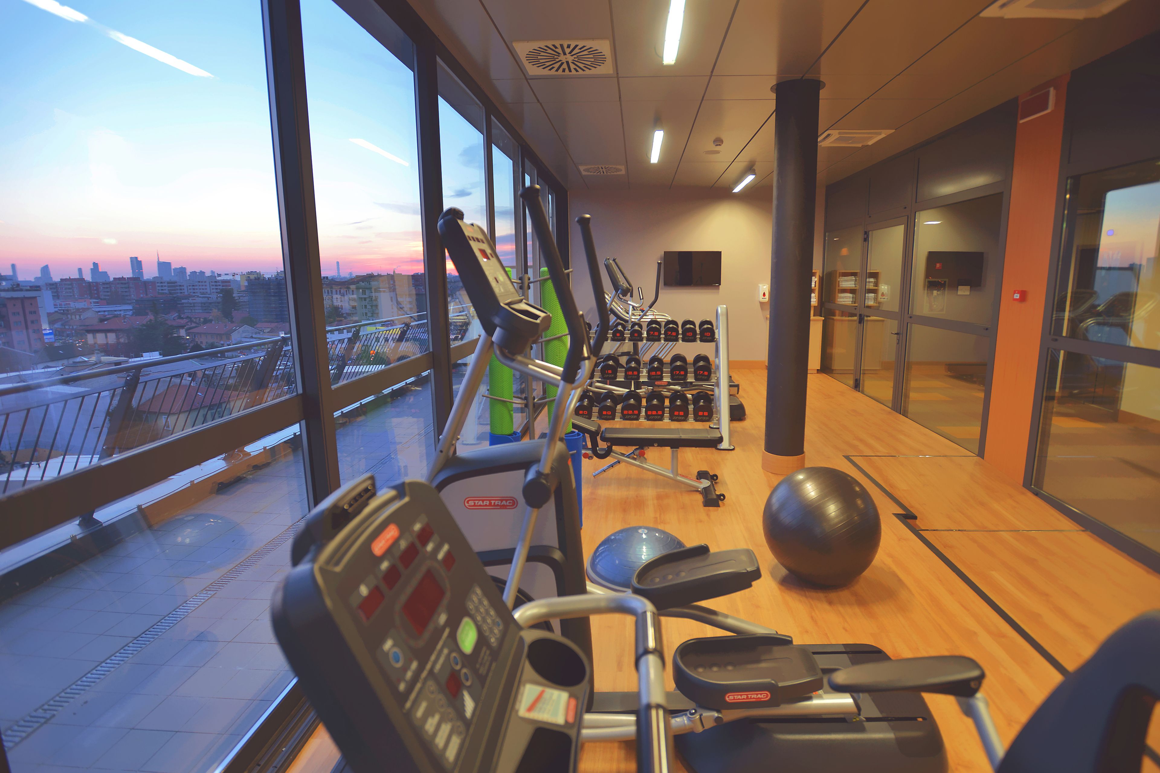 Una sala de gimnàstica amb equip d'exercici, una finestra gran amb vista a la ciutat i un sòl de fusta.