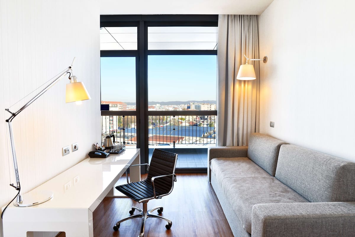 A modern hotel room with a large window, a grey sofa, a desk with a chair, and a lamp.