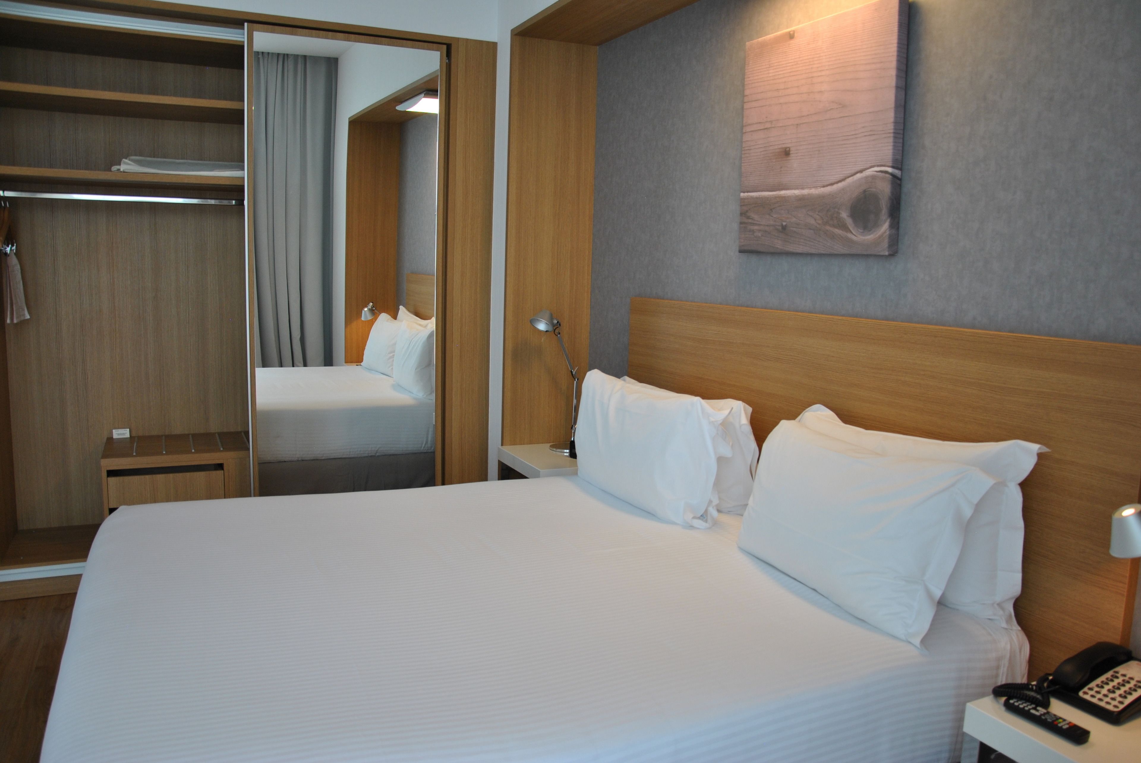 Une chambre d’hôtel avec un grand lit, une tête de lit en bois, une table de chevet avec une lampe et une armoire avec un miroir.