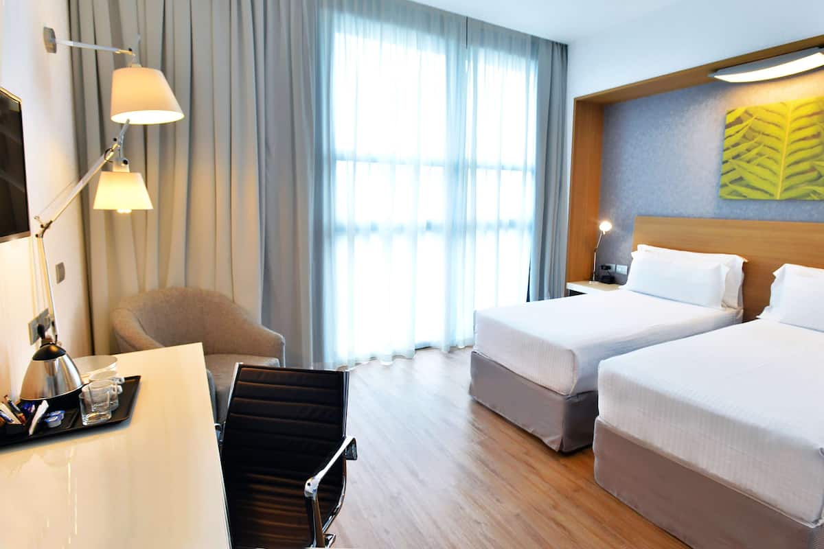 A hotel room with two beds, a desk with a chair, a lamp, and a large window with curtains.