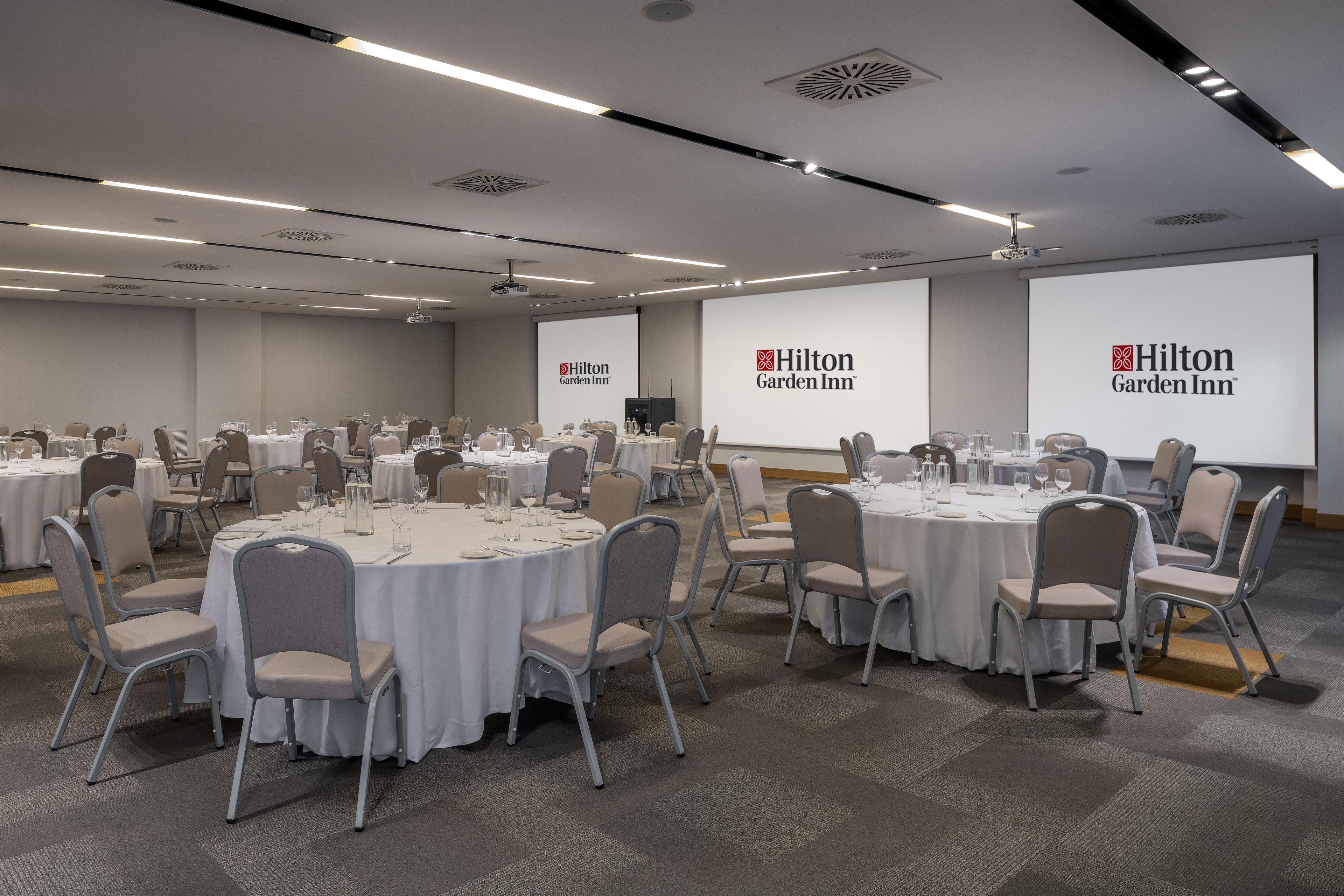 Ein Konferenzraum mit runden Tischen, Stühlen und einer Projektionsleinwand mit dem Logo des Hilton Garden Inn.