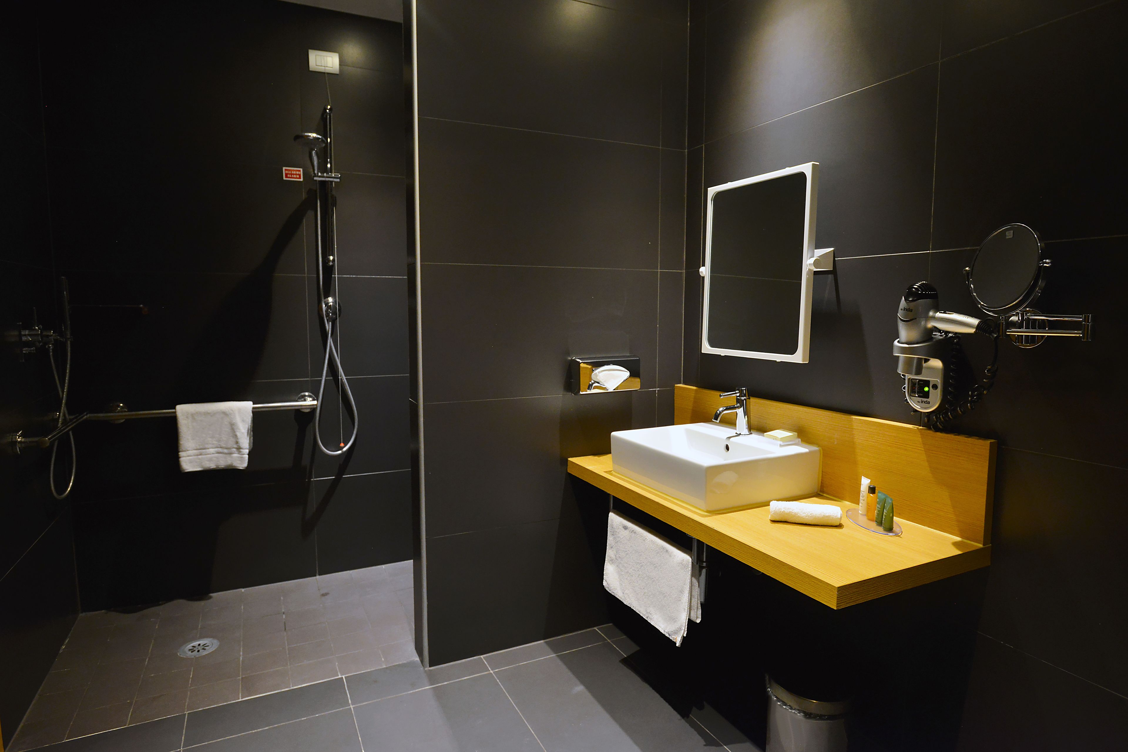 Een moderne badkamer met een donkere betegelde muur, een houten wastafelmeubel met een witte wasbak, een spiegel en een handdouche.