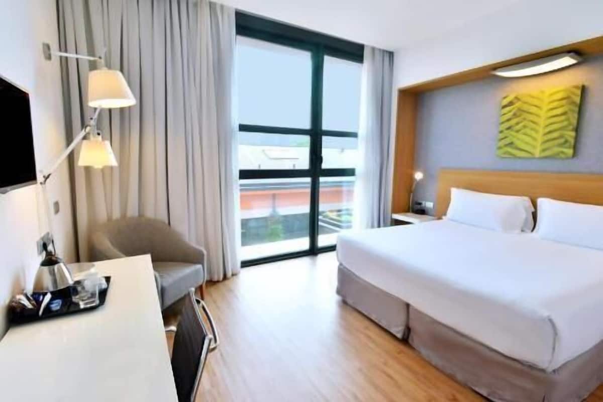 A hotel room with a bed, a desk, a chair, a lamp, and a large window.