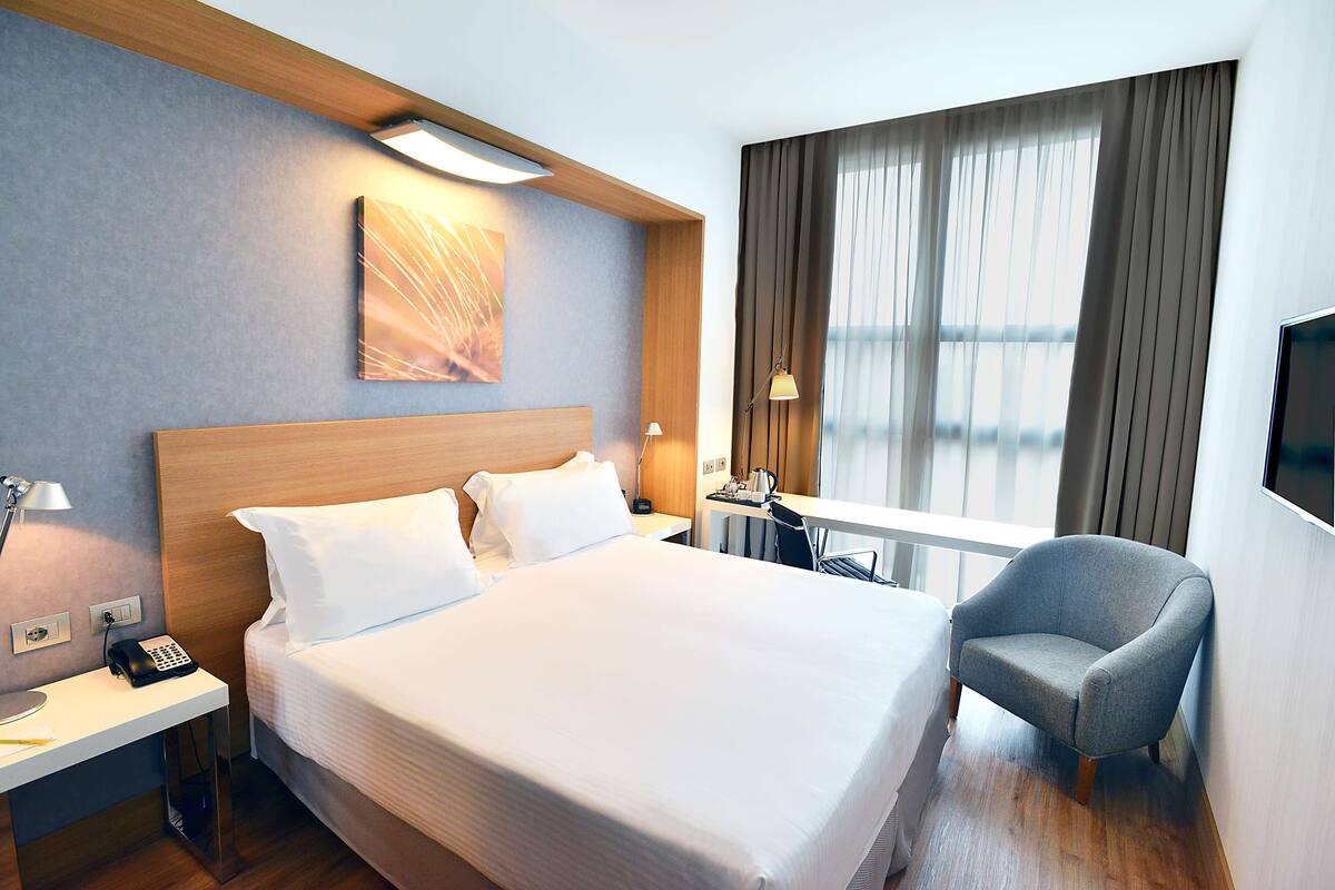 A hotel room with a large bed, a desk with a lamp, a chair, and a TV.