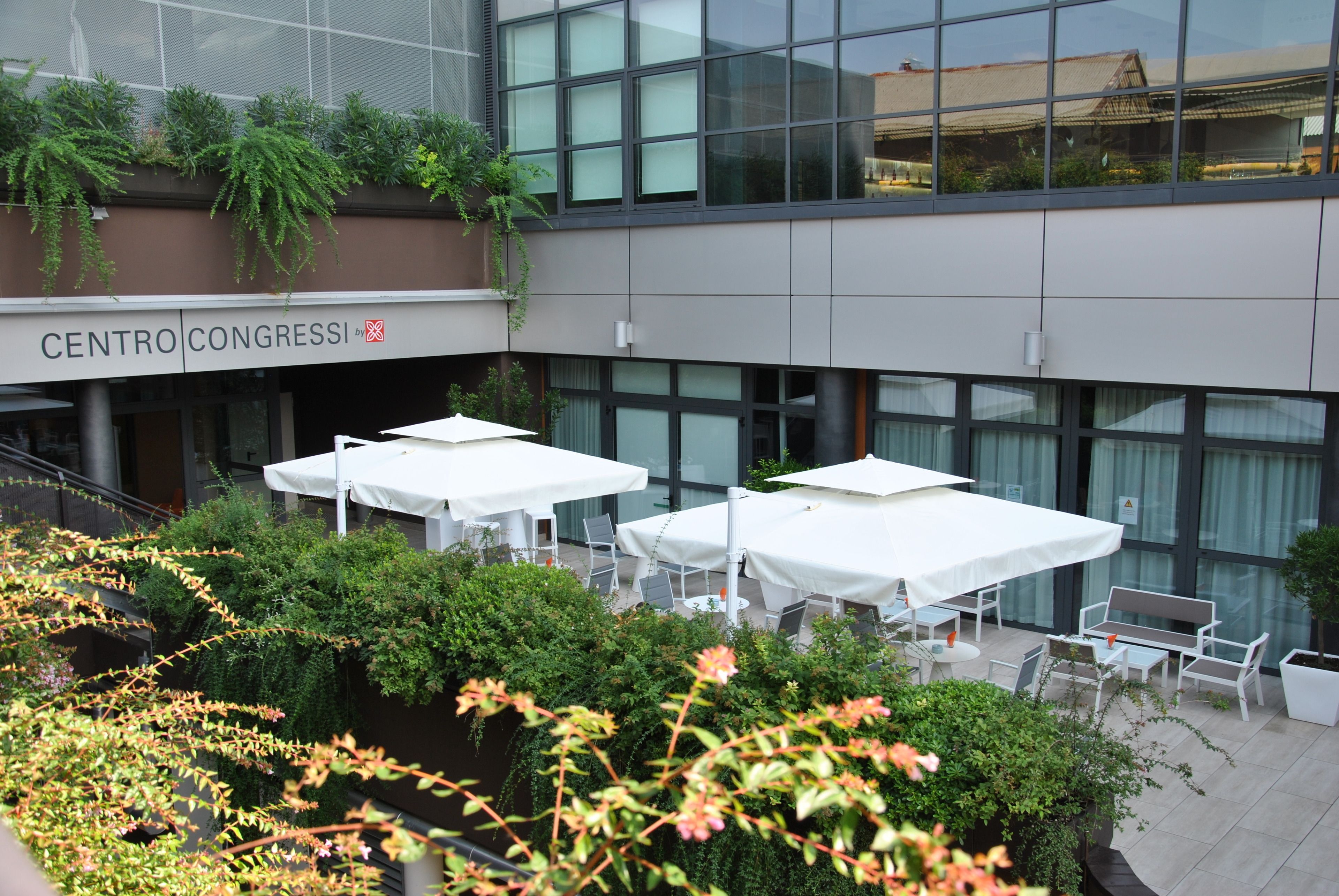 An outdoor seating area with white tables and umbrellas, surrounded by greenery and a building with large windows and a sign reading "CENTRO CONGRESSI".