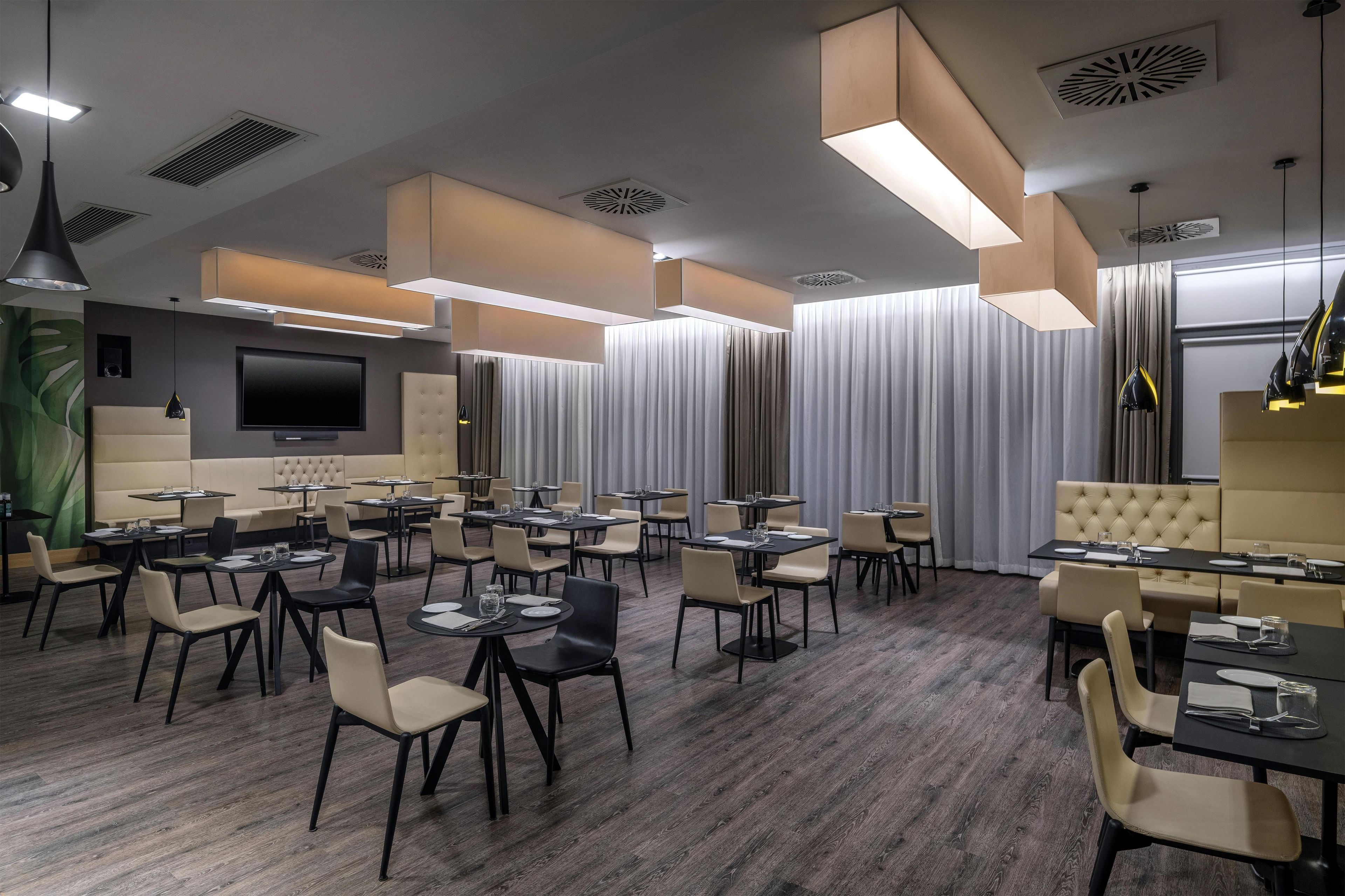 A modern dining area with multiple tables and chairs, a flat-screen TV mounted on the wall, and ceiling vents.