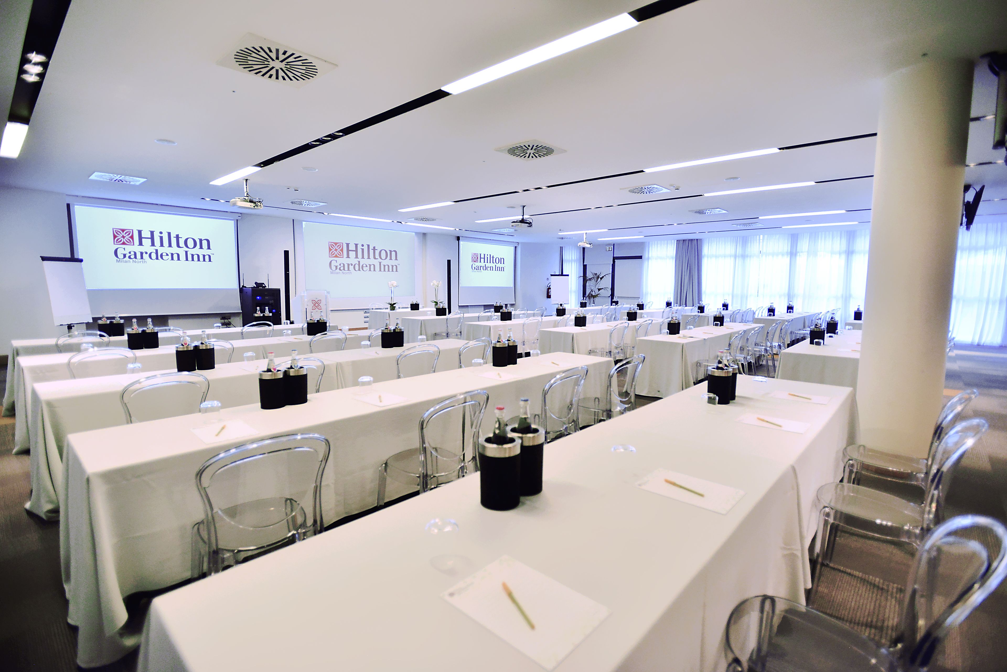 Un salón de conferencias con varias mesas, sillas cómodas y pantallas mostrando el logotipo de Hilton Garden Inn.