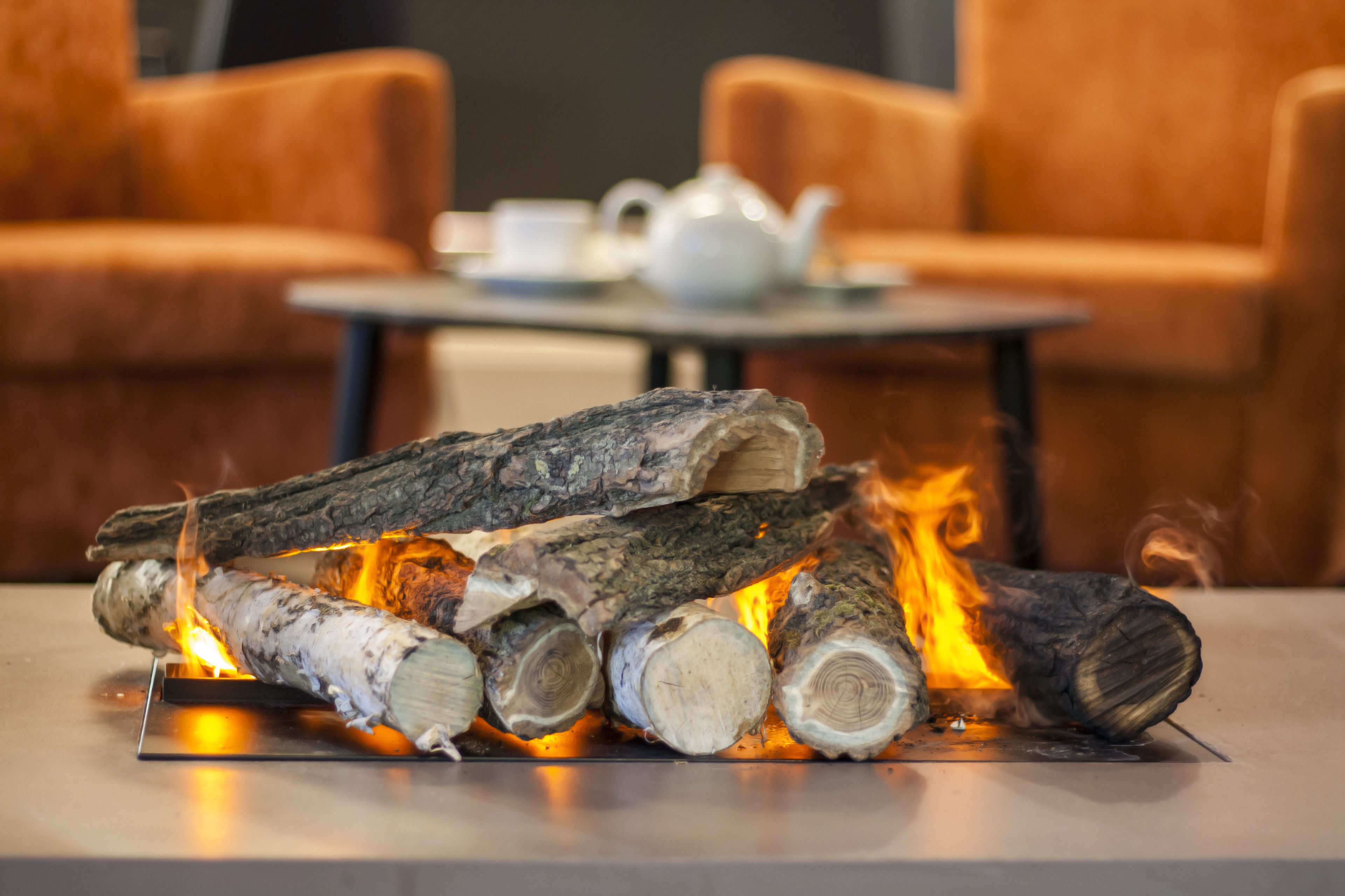 Una chimenea con leña ardiendo, una mesa con tetera, y sillas tapizadas en color naranja al fondo.
