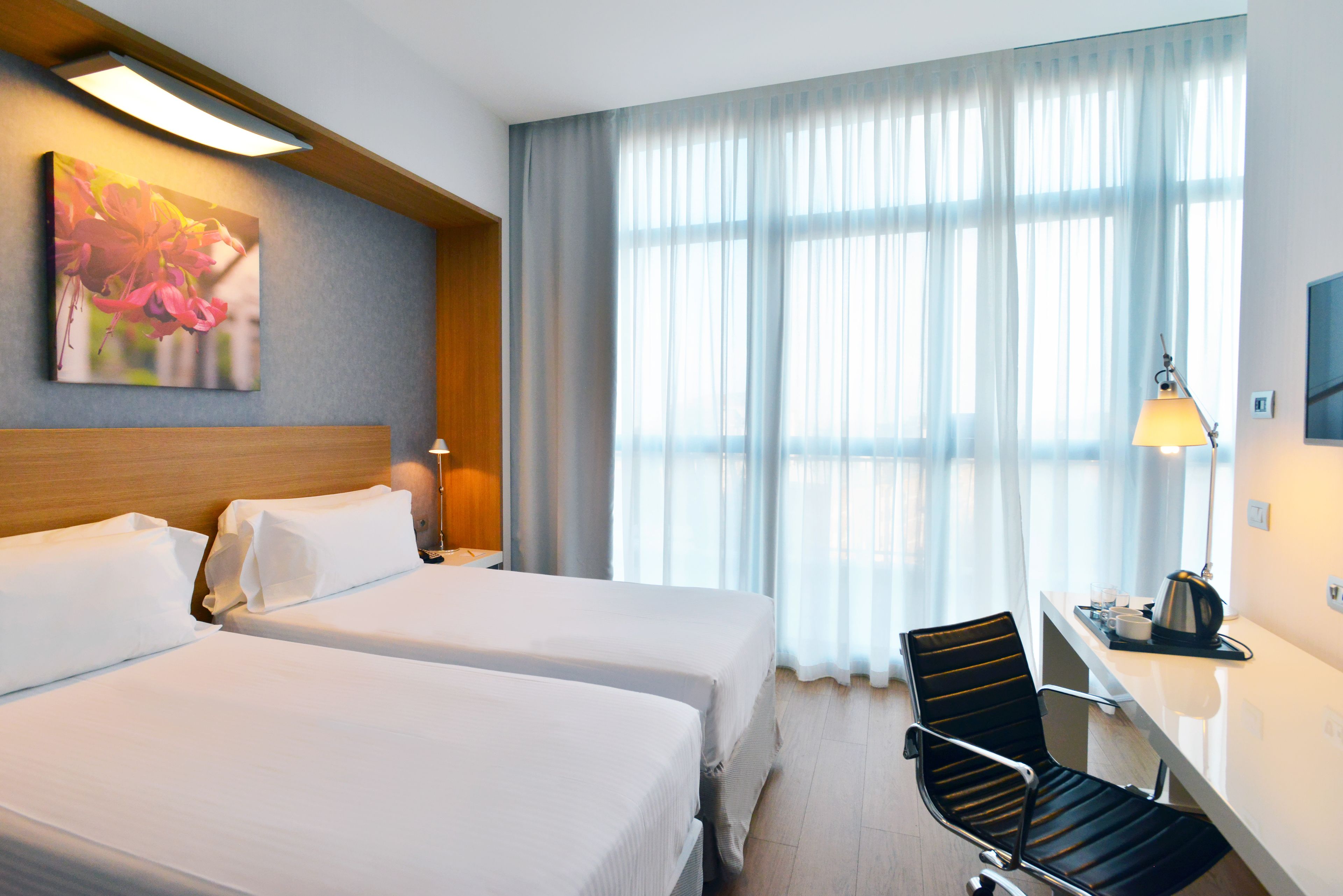 Habitación de hotel con dos camas, un escritorio con silla, una lámpara y un ventanal con cortinas.