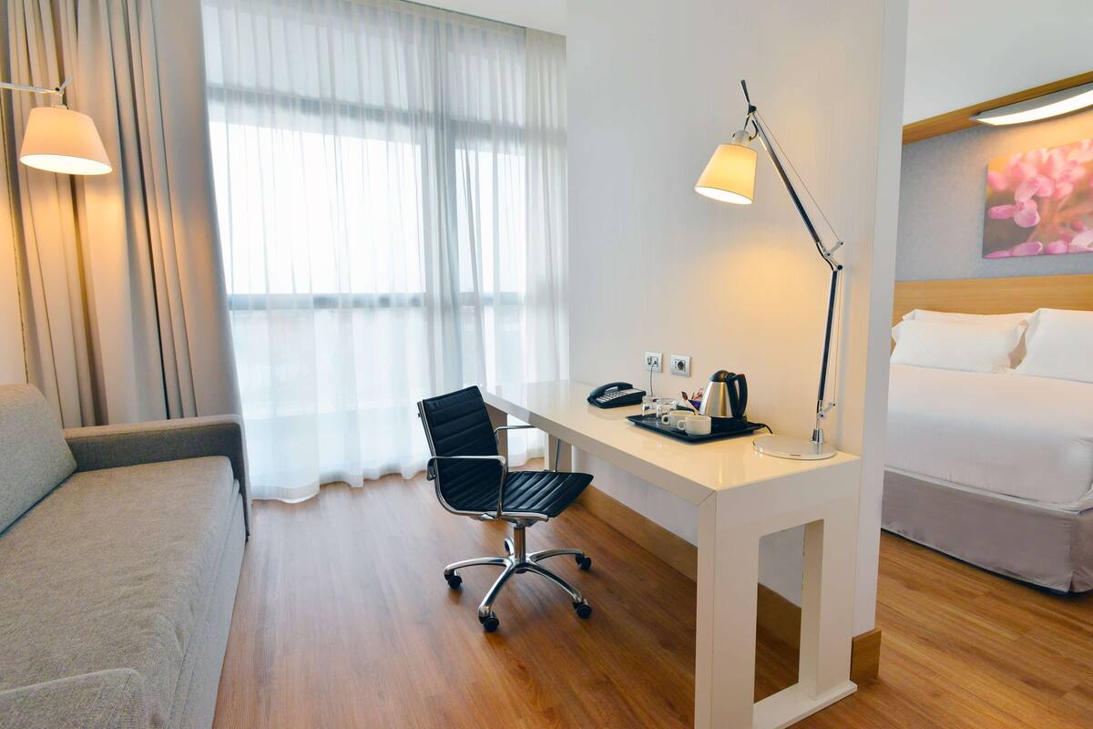 A hotel room with a bed, a desk with a chair, a lamp, and a window with curtains.