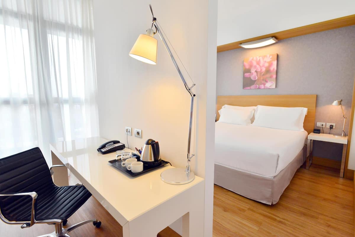 A hotel room with a desk, chair, bed, bedside table, and a painting on the wall.