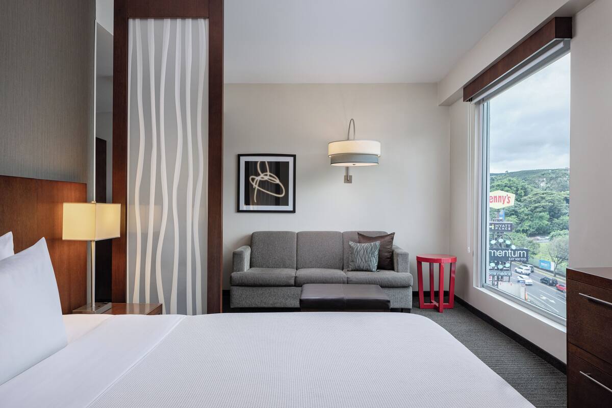 Habitación de hotel con cama, sofá, taburete rojo, lámpara, cuadro enmarco y ventana con vistas.