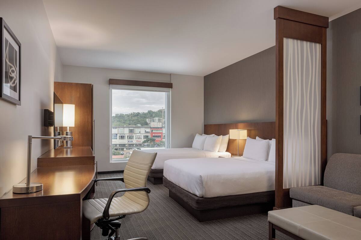 Habitación de hotel con dos camas, un escritorio, una silla, un sofá, una ventana con vistas y una lámpara.
