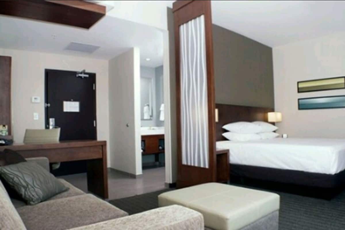 Habitación de hotel con cama, escritorio, silla, sofá y baño visible a través de una puerta abierta.