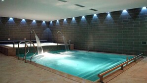 An indoor swimming pool with a waterfall feature, surrounded by a brick wall and equipped with metal railings. - Bellaria (Predazzo)