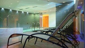 A modern indoor pool area with clear glass walls, a central water feature, and a staircase with metal railings. - Bellaria (Predazzo)