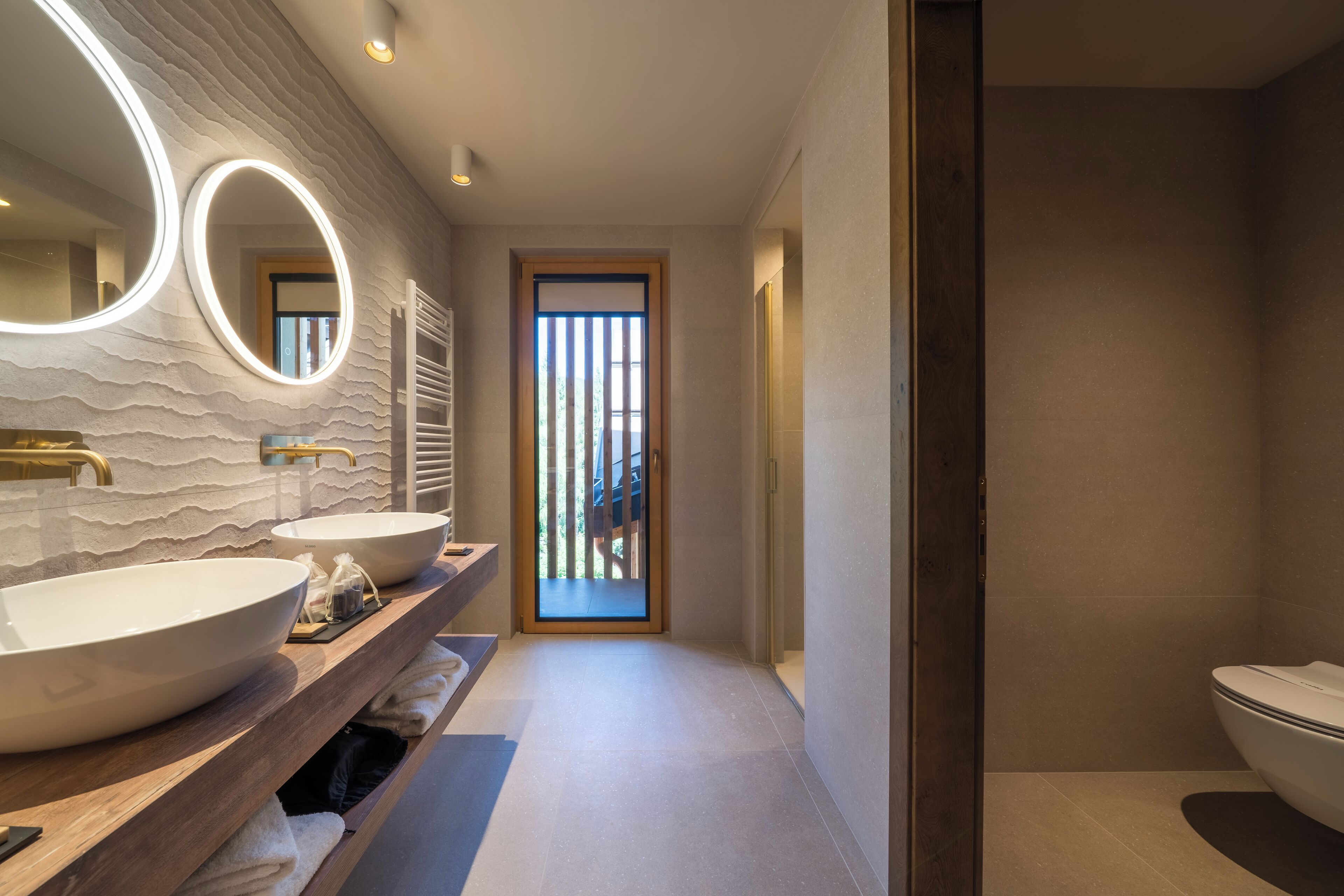 Une salle de bain moderne avec deux grands lavabos, un meuble-lavabo en bois et un grand miroir agrémenté de lumières disposées en cercle.