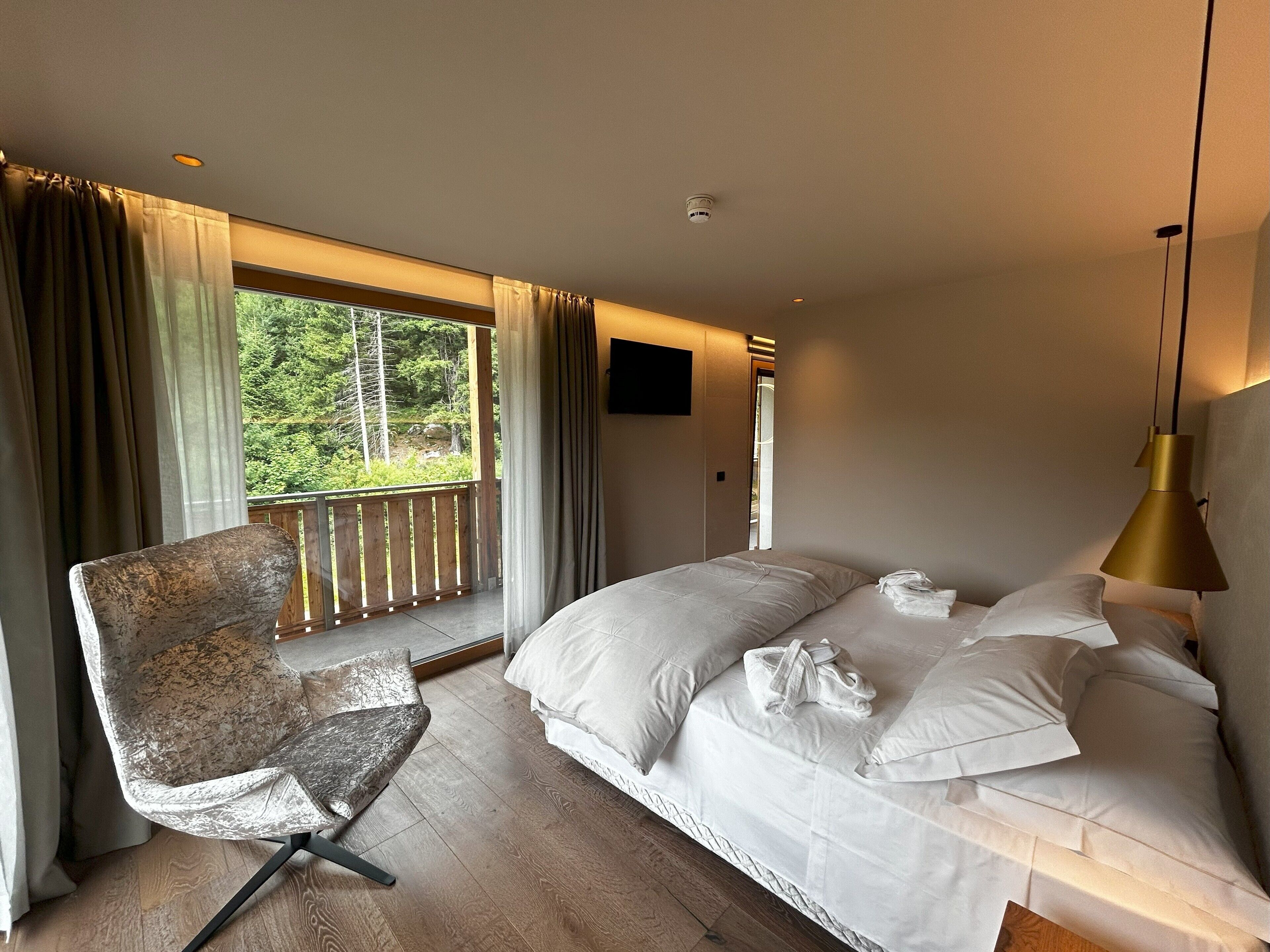 Een slaapkamer met een groot bed, een stoel, een balkon met uitzicht op bomen en een plafondlamp.