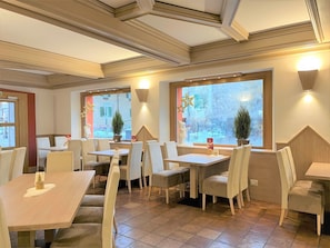Un interior de restaurante con varias mesas y sillas, ventanas amplias con detalles decorativos y un piso de baldosas.