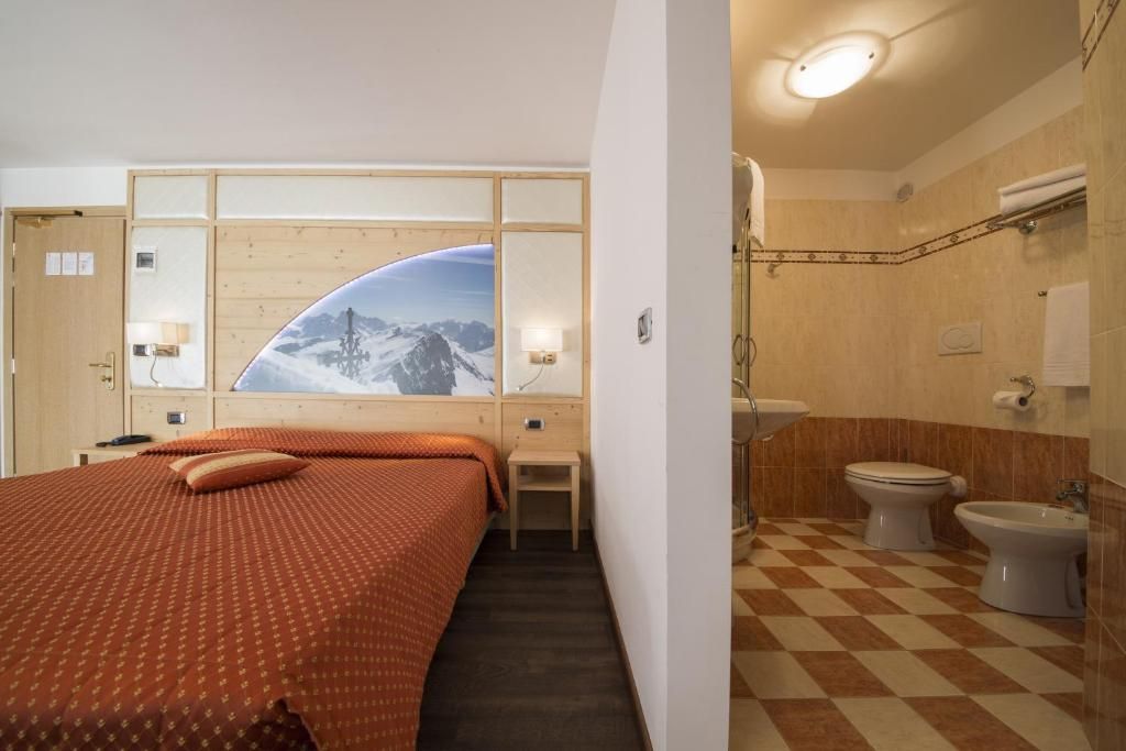 Habitación de hotel con cama, una mesita, un espejo en la pared, ventana con vista a montañas y baño con inodoro, bidé y piso a cuadros.