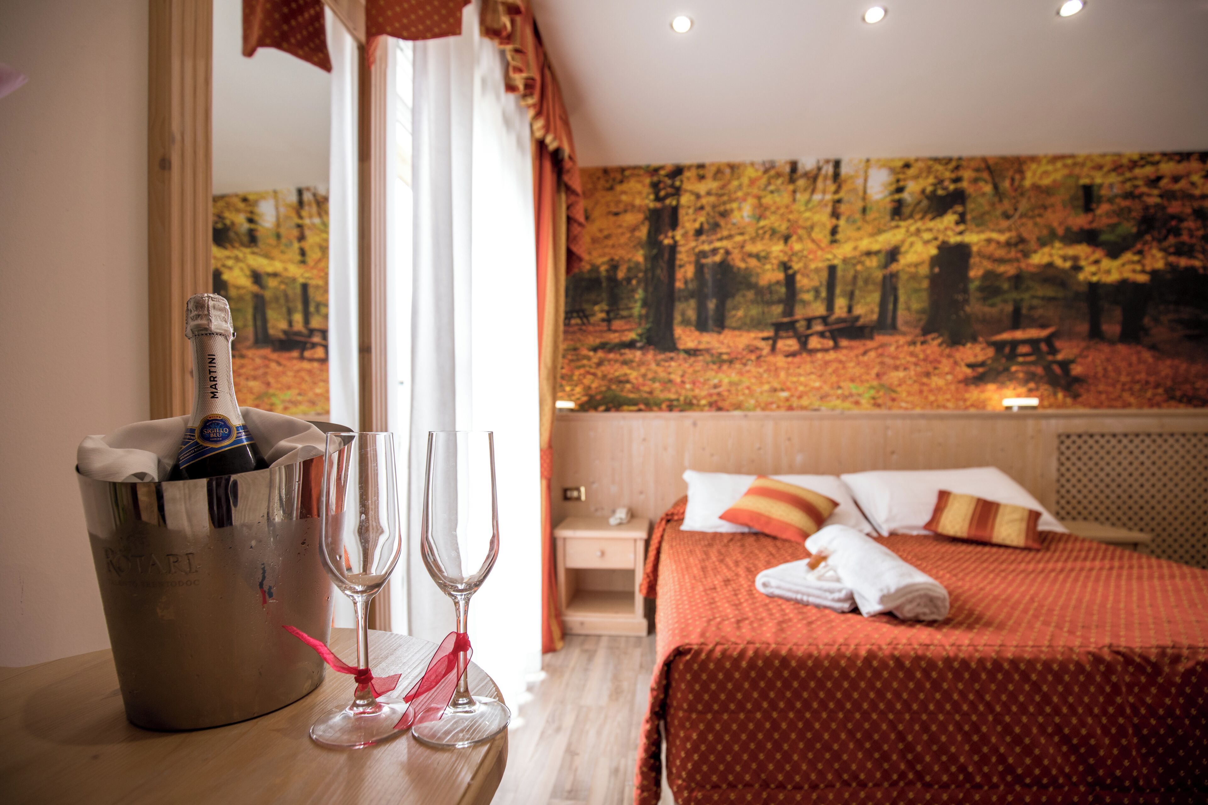 Une chambre d’hôtel avec un lit, une table de chevet, un tableau représentant une forêt, un minibar et deux verres.