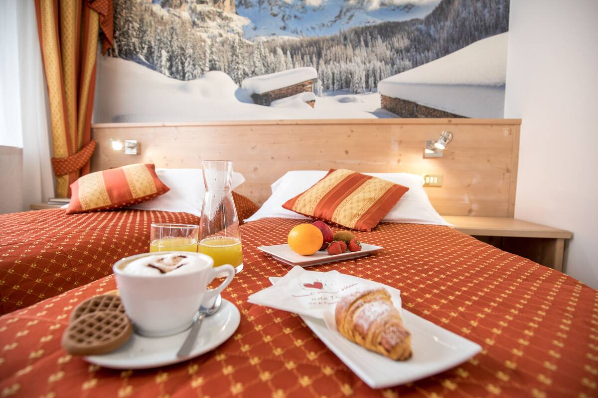 A hotel room with a bed, breakfast setup including croissant, orange, and strawberries, and a glass of orange juice.