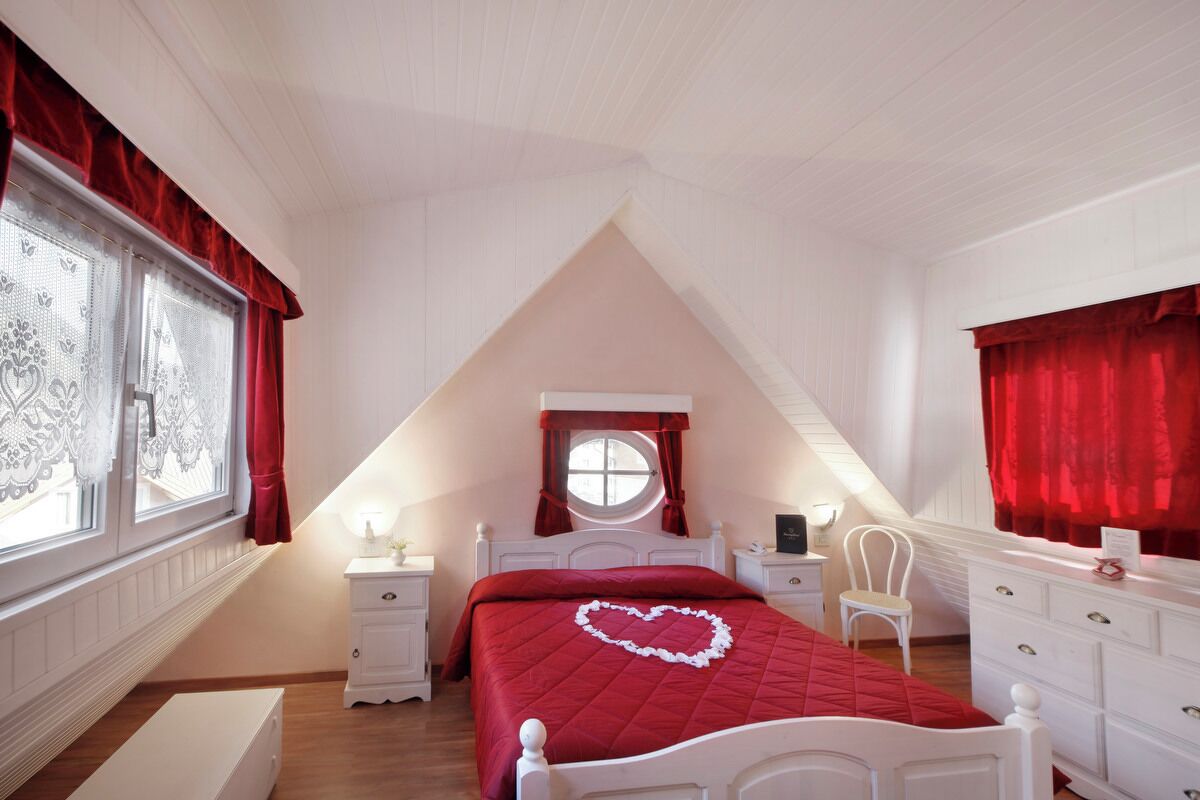 A bedroom with a sloped ceiling, a bed with a heart-shaped decoration, a window with lace curtains, and a dresser.