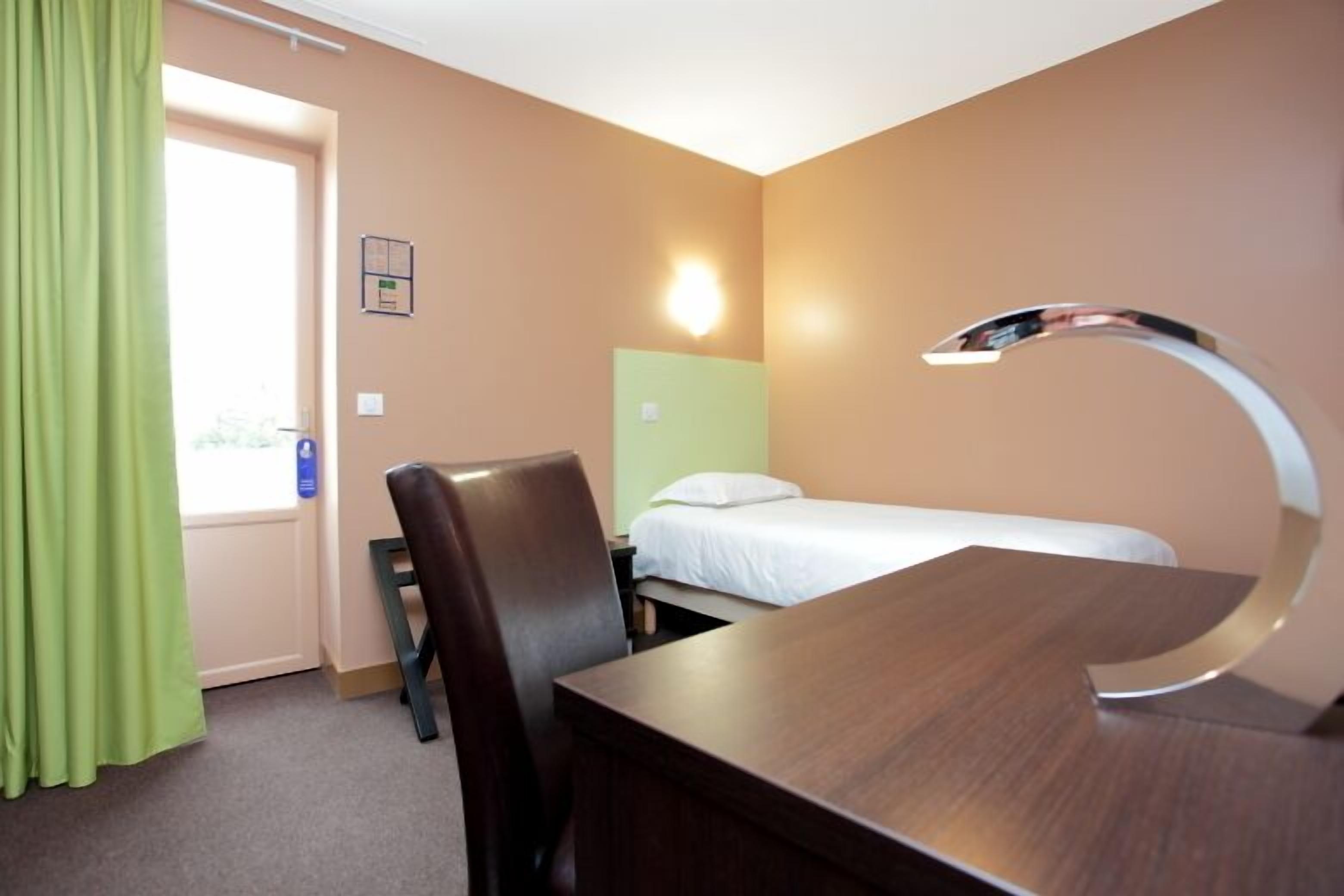 A hotel room with a bed, a desk, a chair, a bathroom mirror, and a window with curtains.